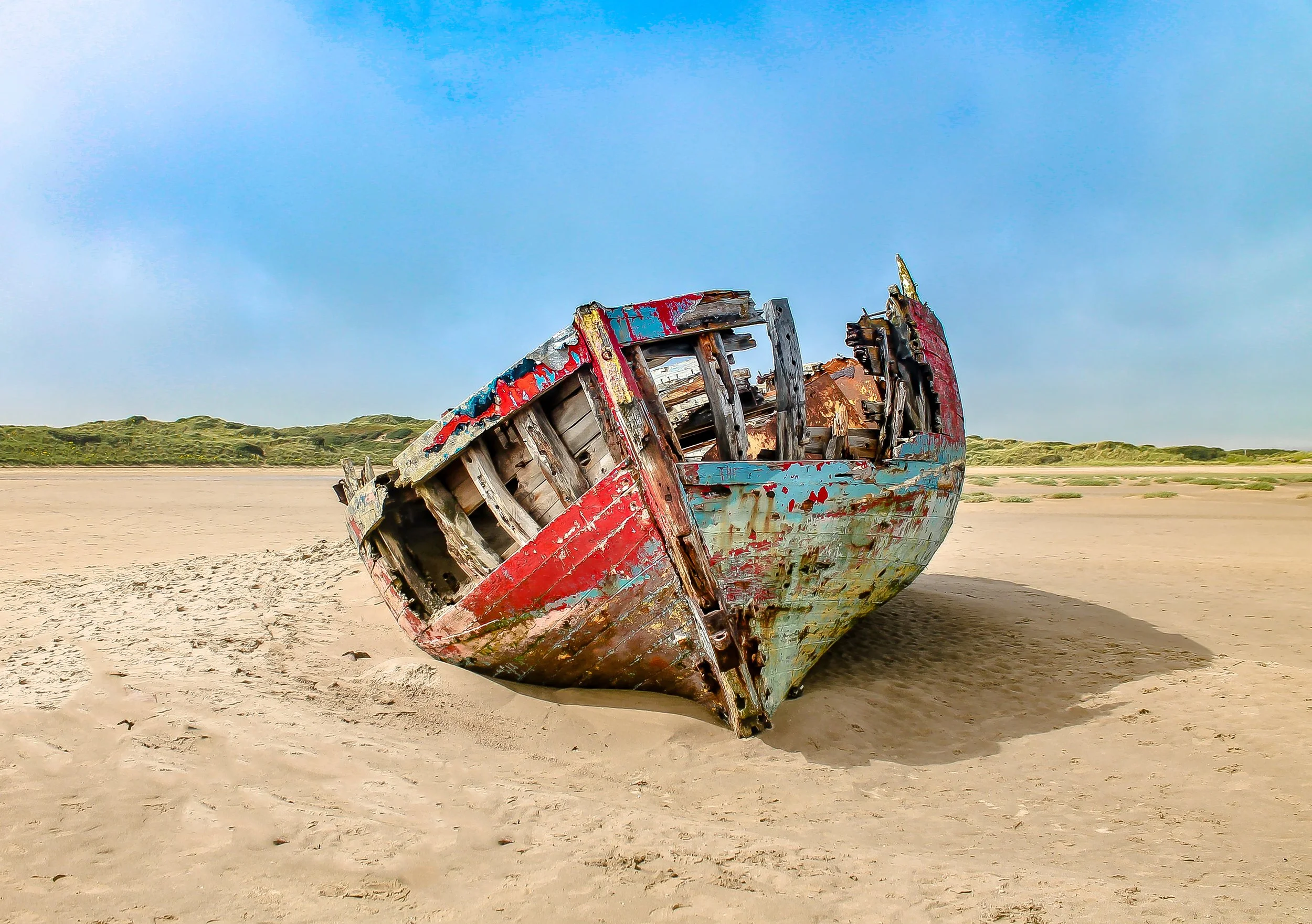 Photo by Kim Stone of abandoned, weathered boat resting on a sandy beach with sparse green vegetation in the background and a partly cloudy blue sky above.