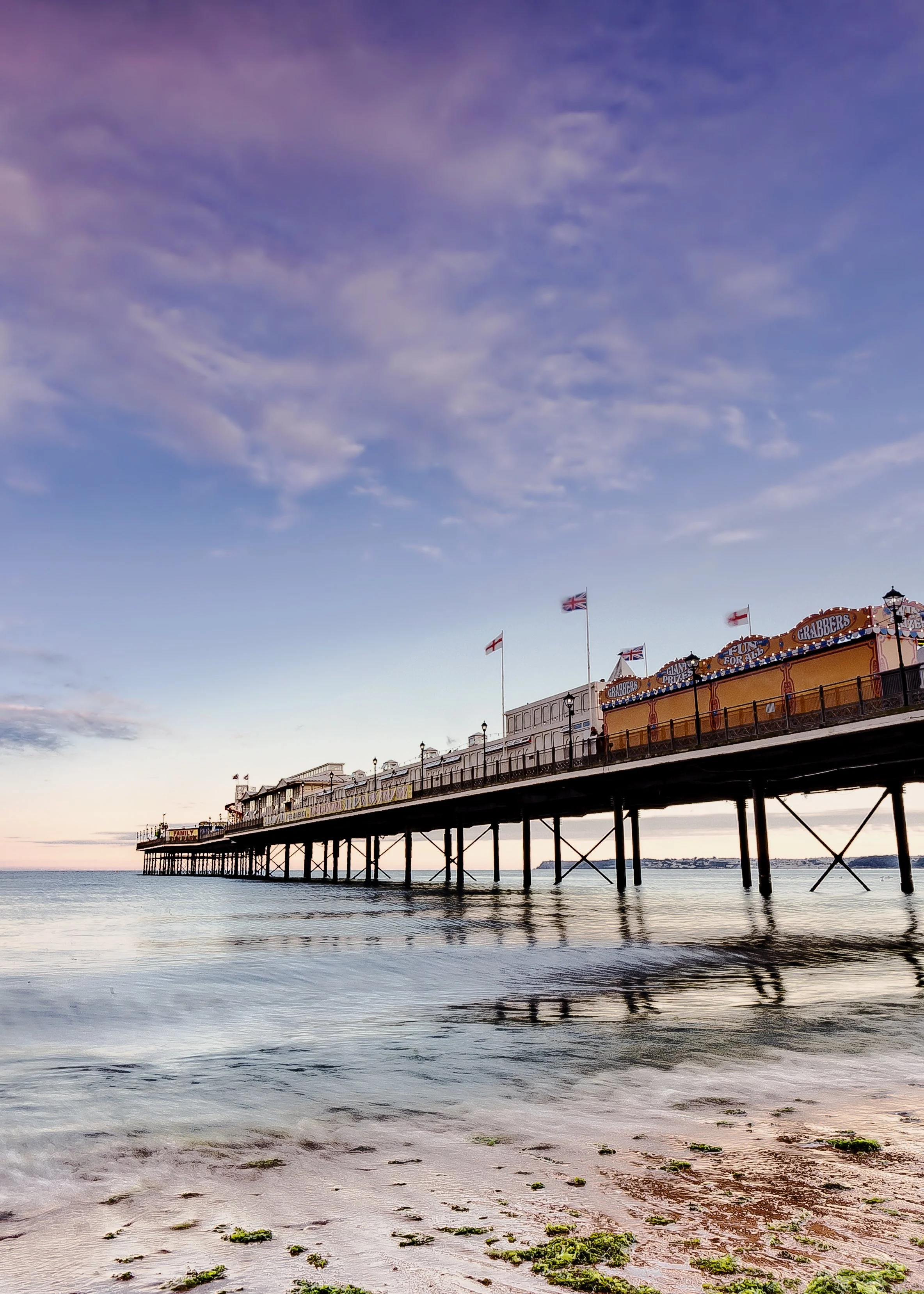 Photo by Kim Stone of Teignmouth Pier extending into the ocean with flags flying on it during sunset or sunrise, calm water below, and a partly cloudy sky overhead.