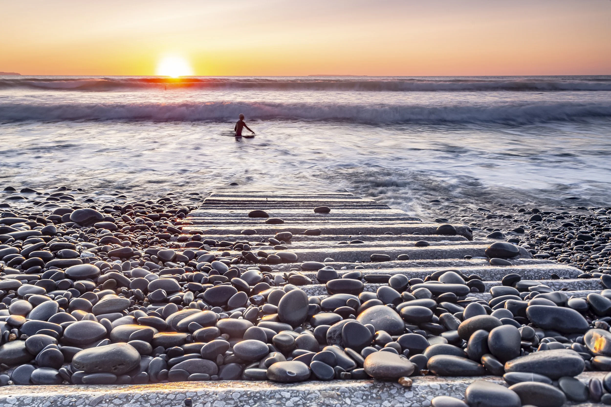 Photo by Kim Stone in Westward Ho! A person surfing in the ocean at sunset, viewed from a pebble beach with a concrete pier leading into the water.