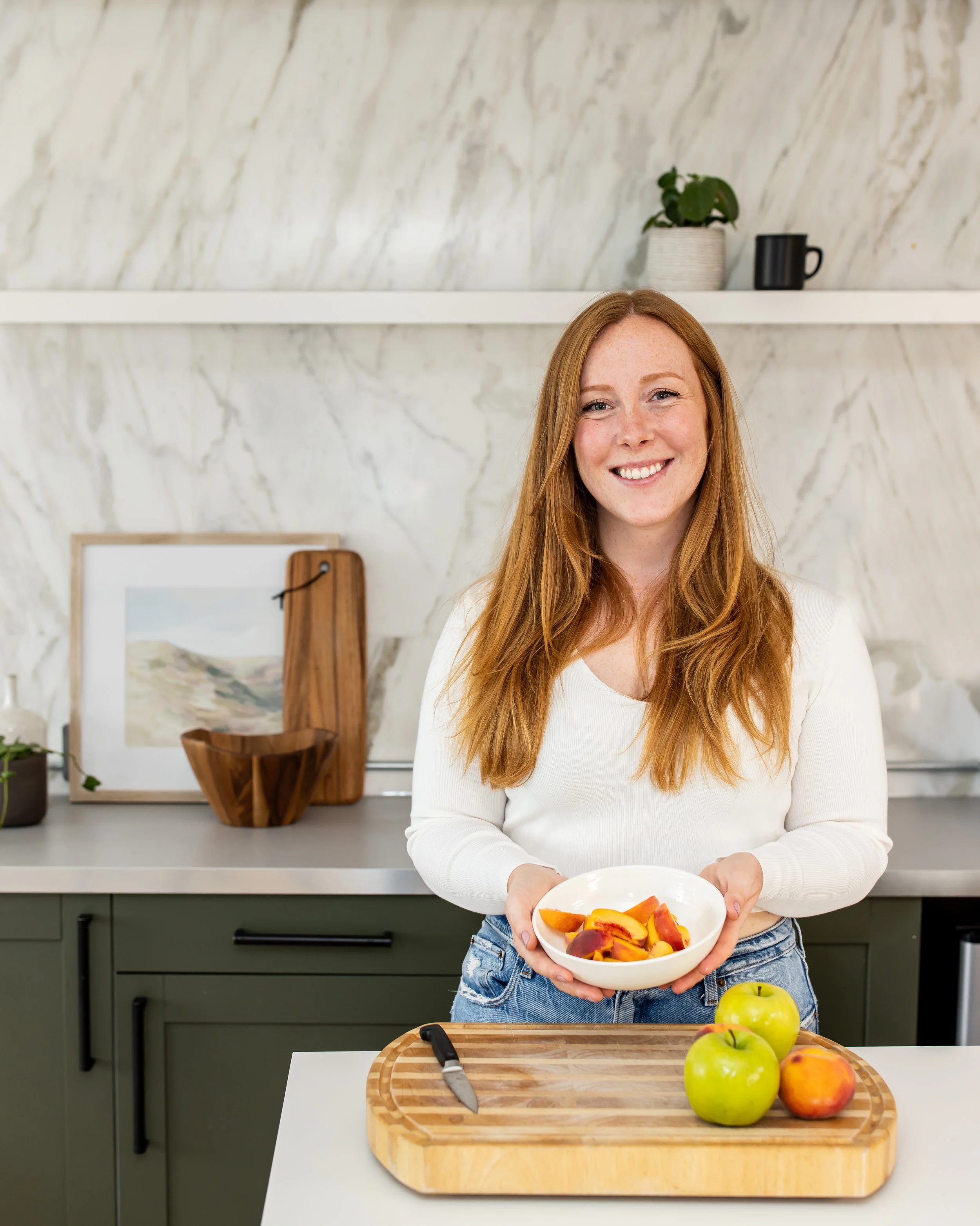 A young woman with red hair and a white sweater smiling and holding a bowl of sliced peaches in a kitchen with a marble wall, green cabinets, and a wooden cutting board with three green apples and a peach.