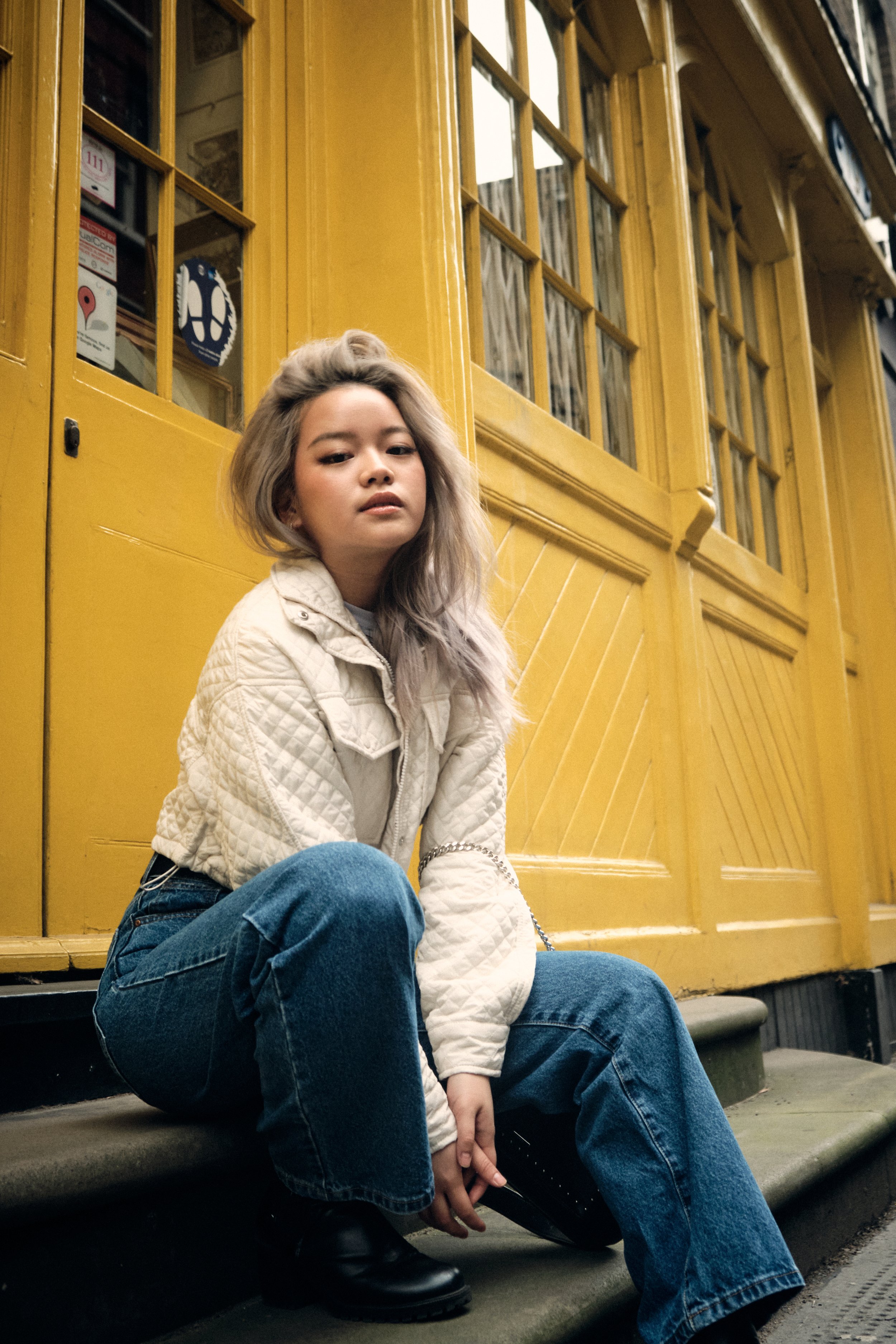 Person with blonde hair sitting on steps, wearing a white quilted jacket and blue jeans, in front of a yellow building with glass paneled doors.