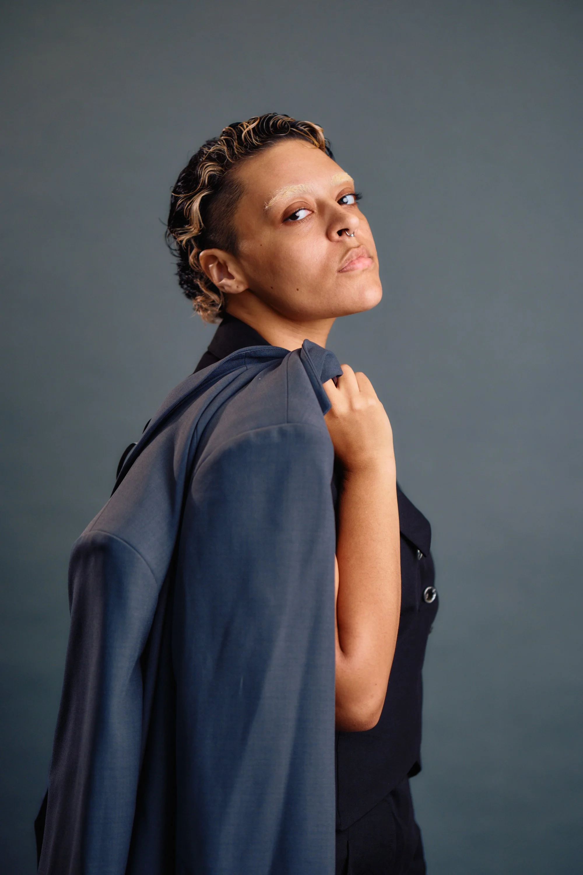 A person with short, curly hair and a nose piercing holding a jacket over their shoulder, looking confidently at the camera.