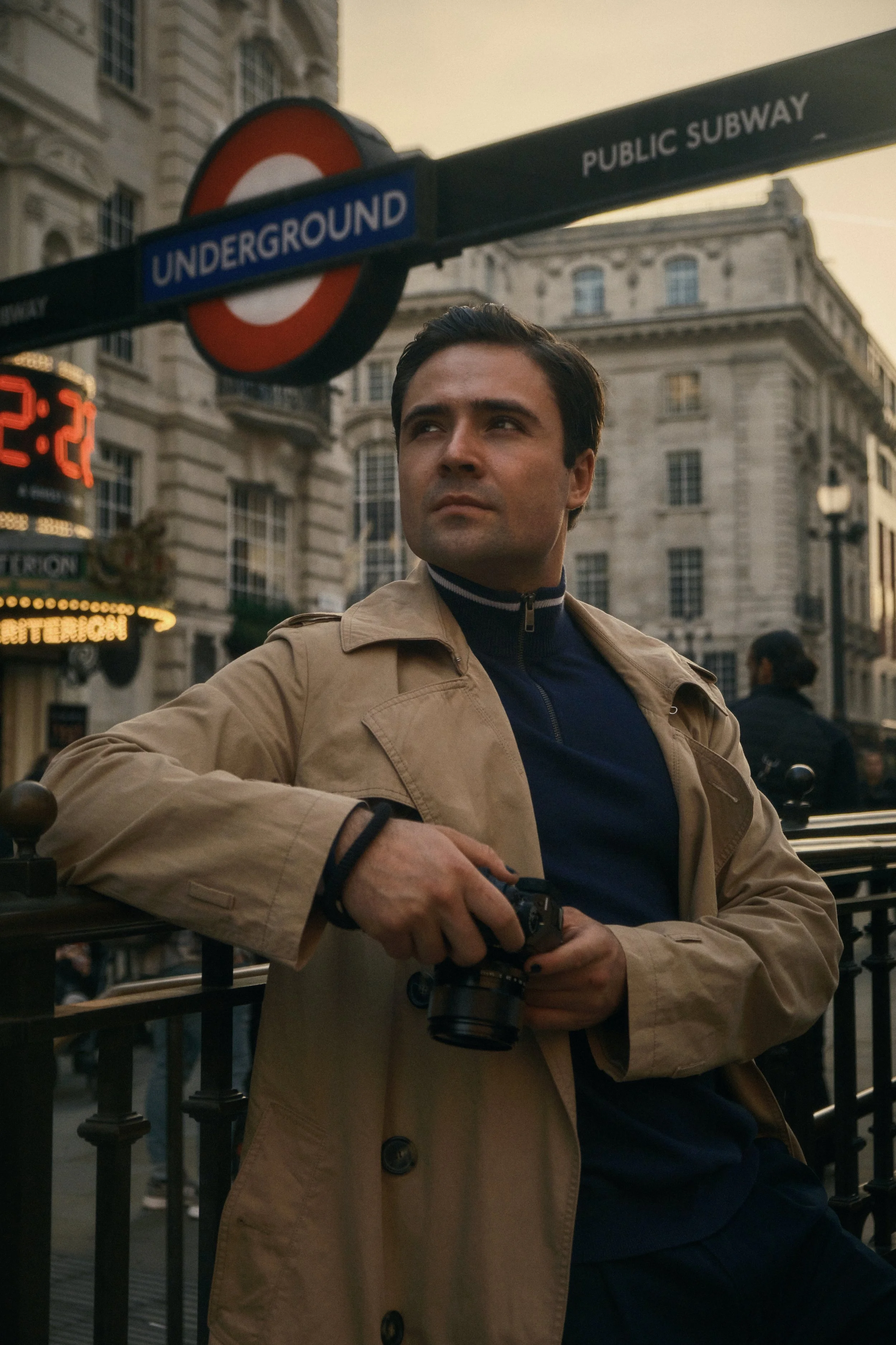 Man in a tan trench coat holding a camera near an Underground subway sign with historical buildings in the background.