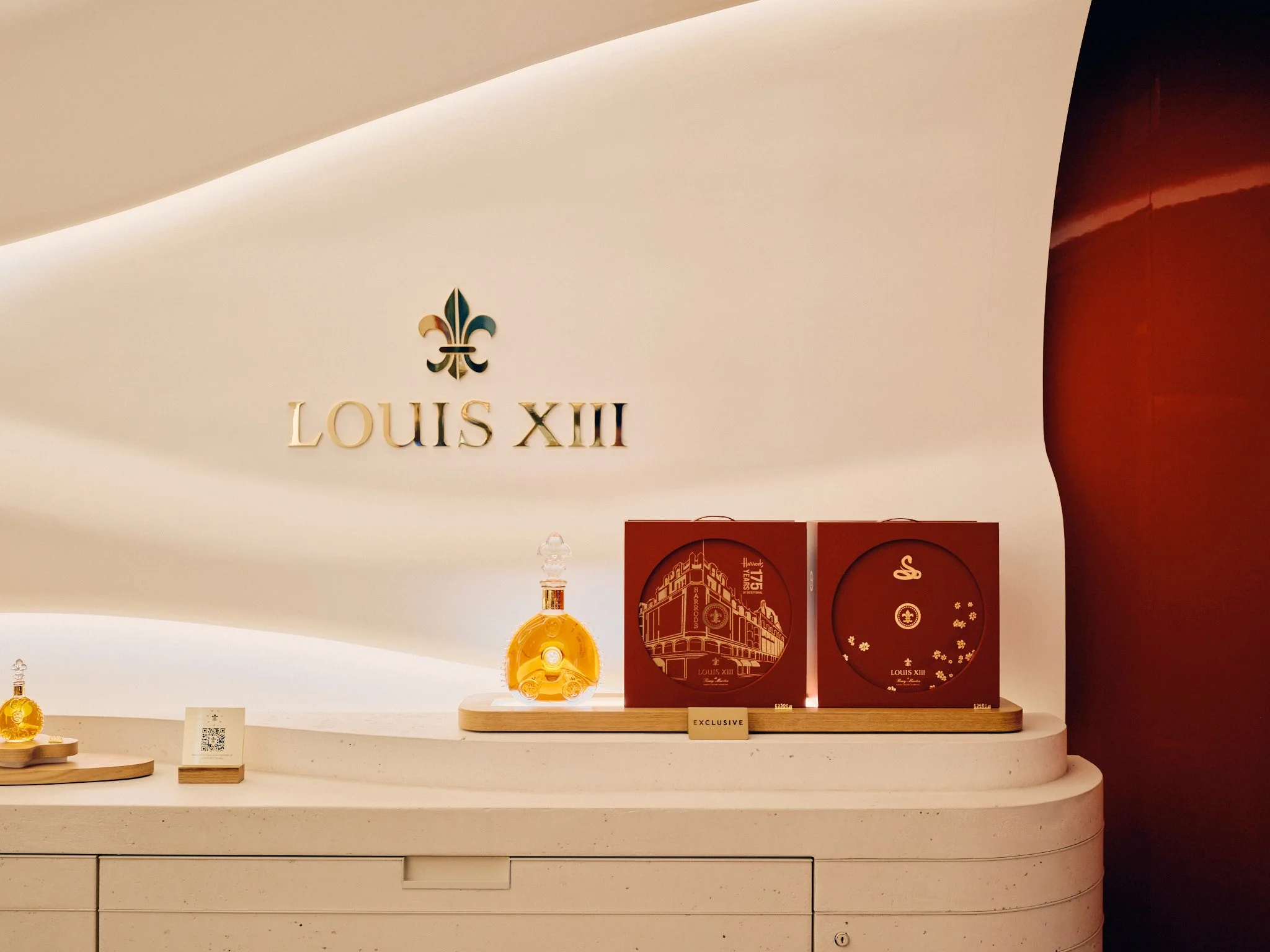 Display of Remy Martin - Louis XIII - Cognac products, including a decorative bottle and packaging, on a white curved surface with Louis XIII logo and fleur-de-lis symbol on the wall.