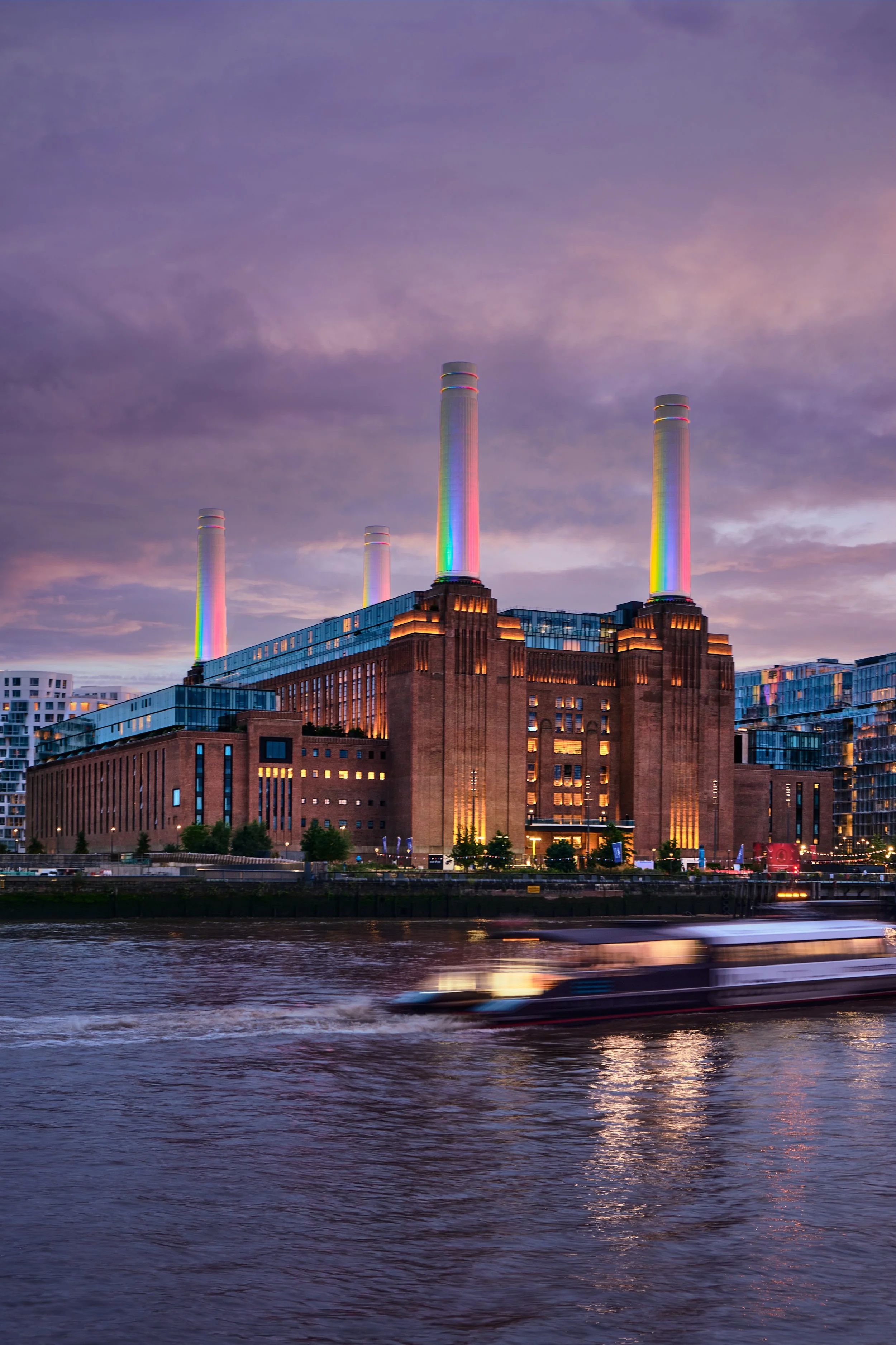 Battersea Power Station, A large historic brick building with four tall chimneys, illuminated with colorful lights, near a river at dusk. A boat is moving across the water in front of the building.