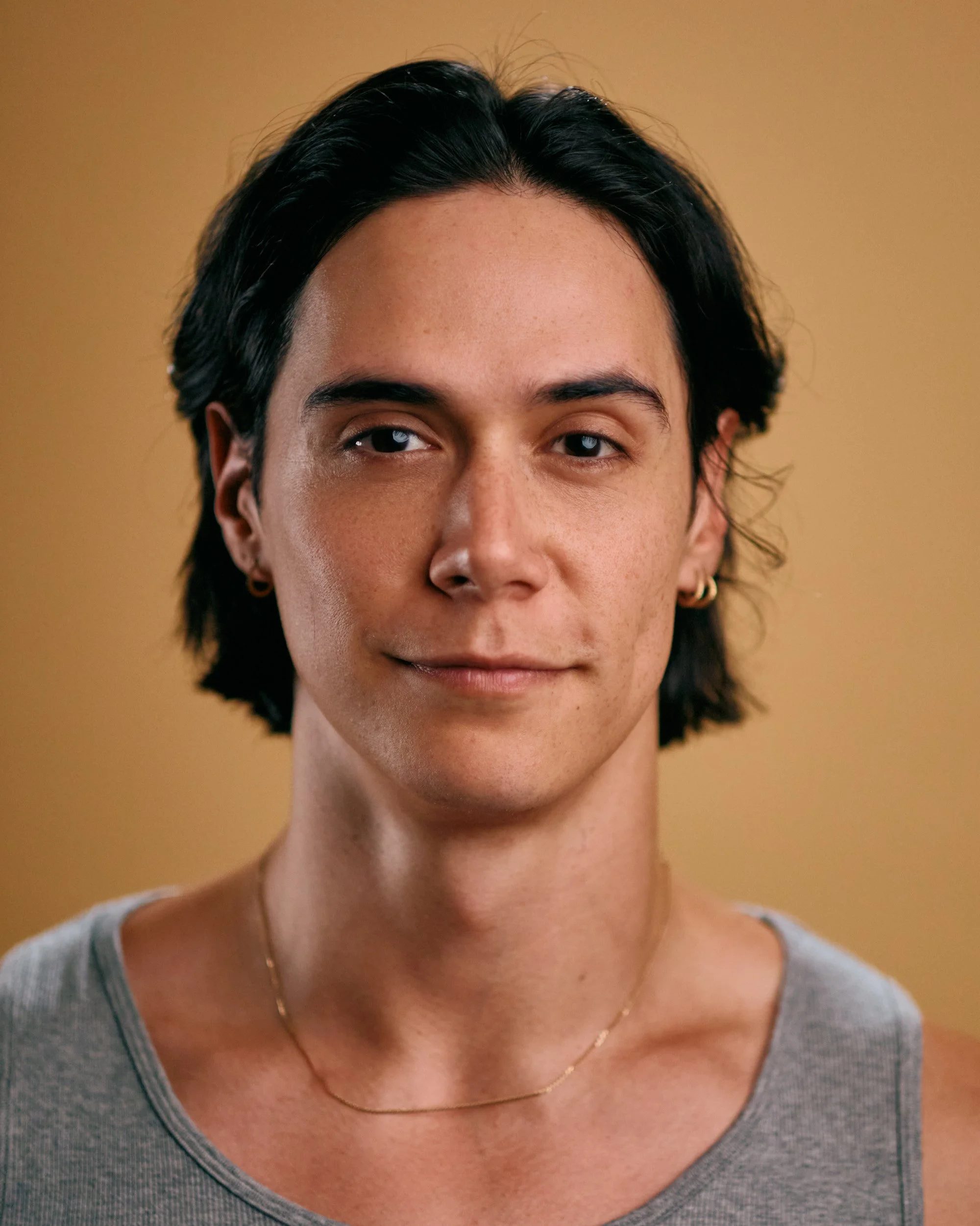 Close-up portrait of a person with dark, shoulder-length hair, fair skin, small earrings, and a thin gold necklace, wearing a gray sleeveless top, against a beige background.