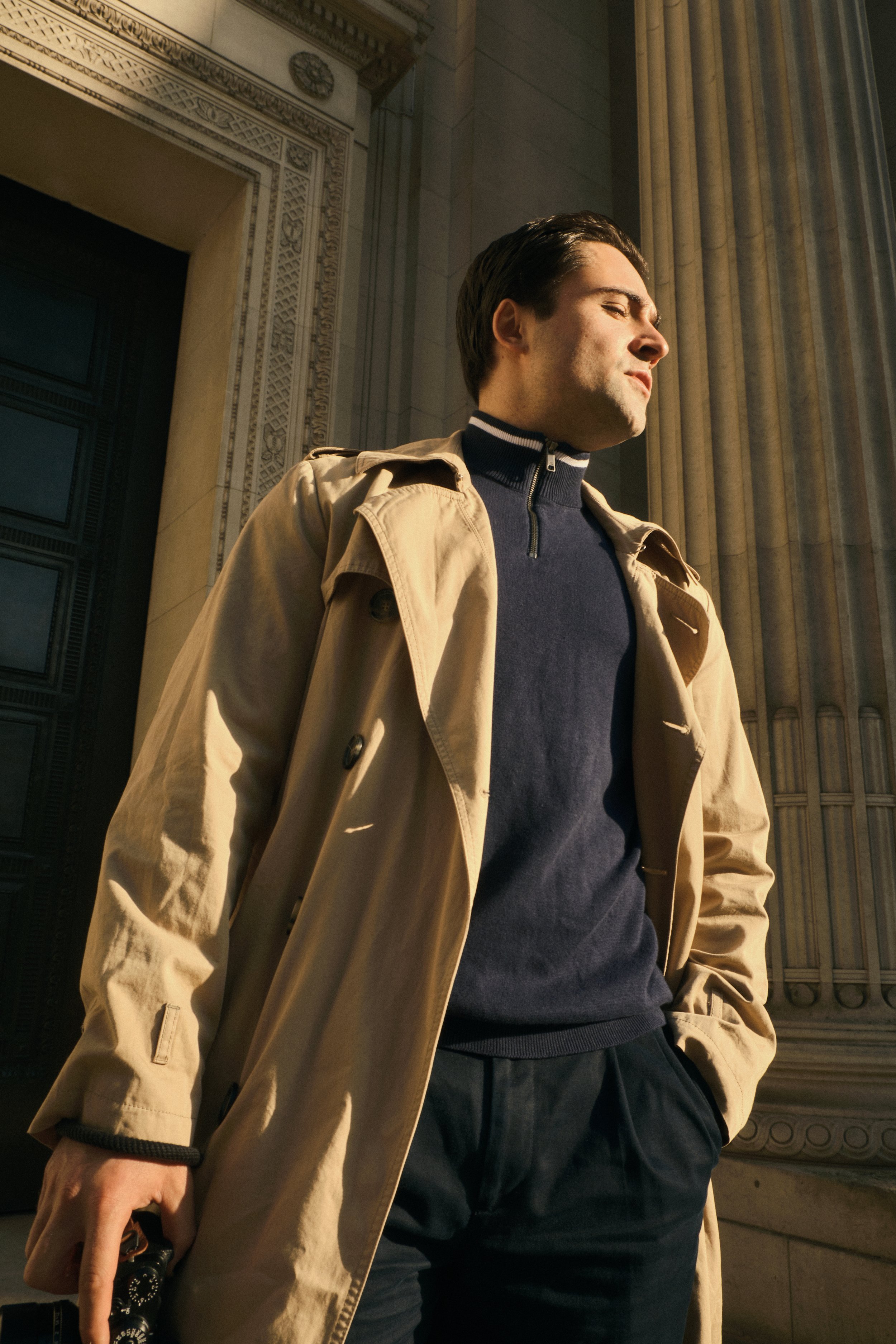 Person wearing a trench coat standing beside a columned building.