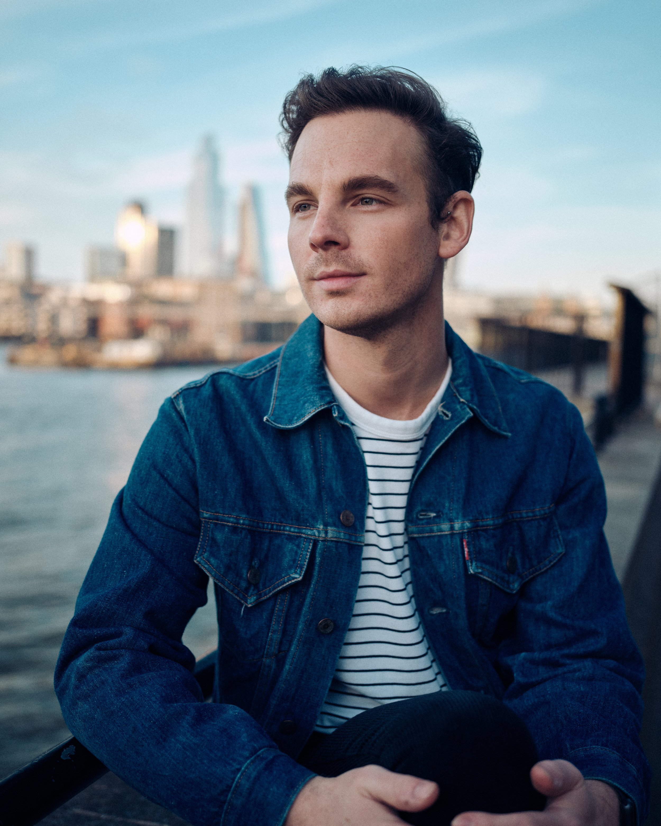 Man in denim jacket sitting near water with cityscape background, clear sky