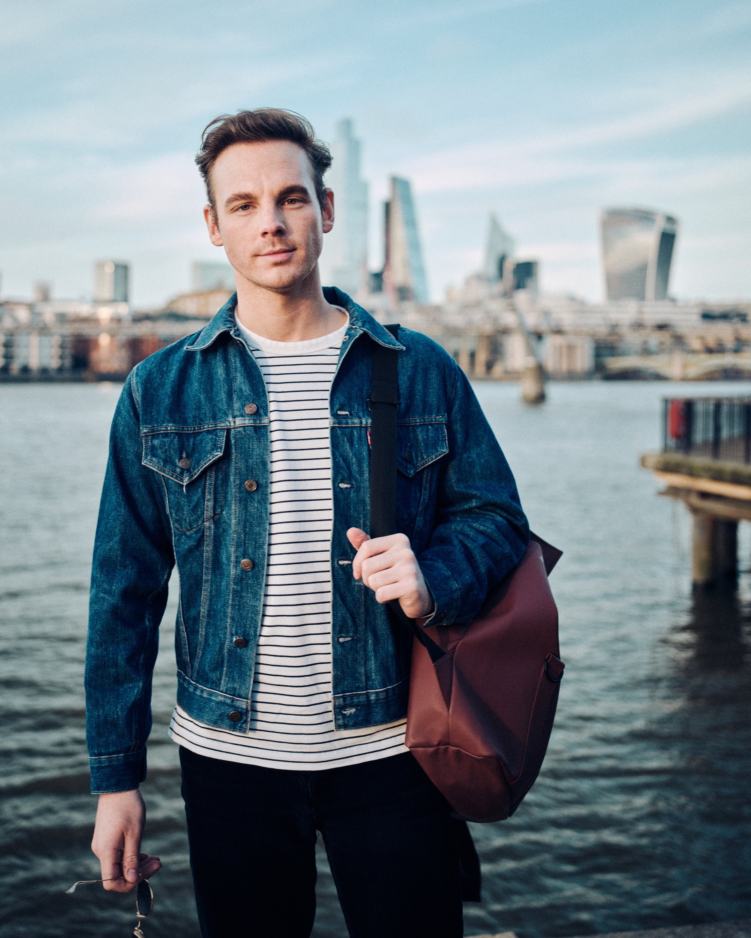 Man in a denim jacket and striped shirt standing by a river with city skyline in the background.