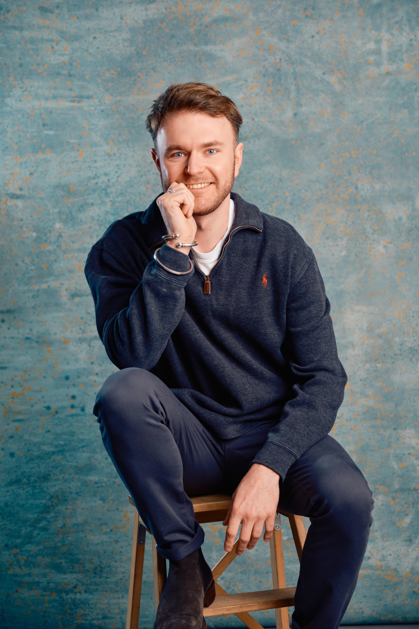 Man sitting on a stool, wearing a dark sweater and smiling with one hand under his chin, blue textured background.