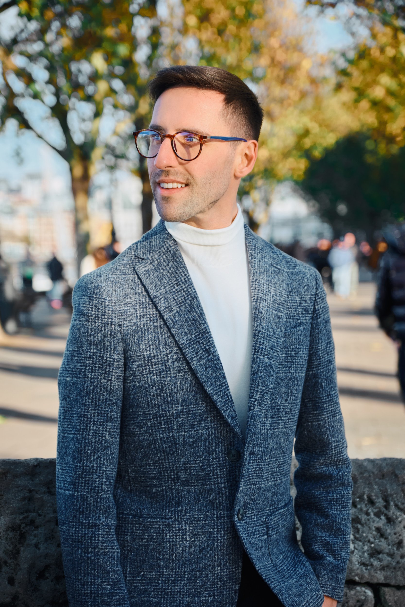Man wearing glasses and a gray blazer standing in an outdoor setting with trees.