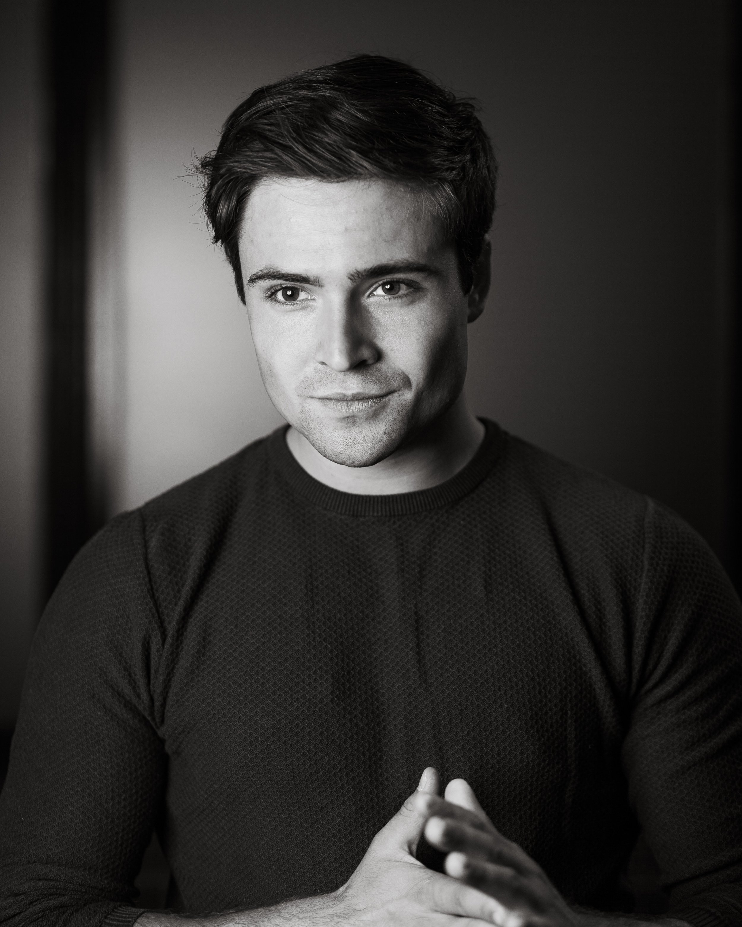 Black and white portrait of a man with short hair, wearing a textured sweater and clasping his hands.