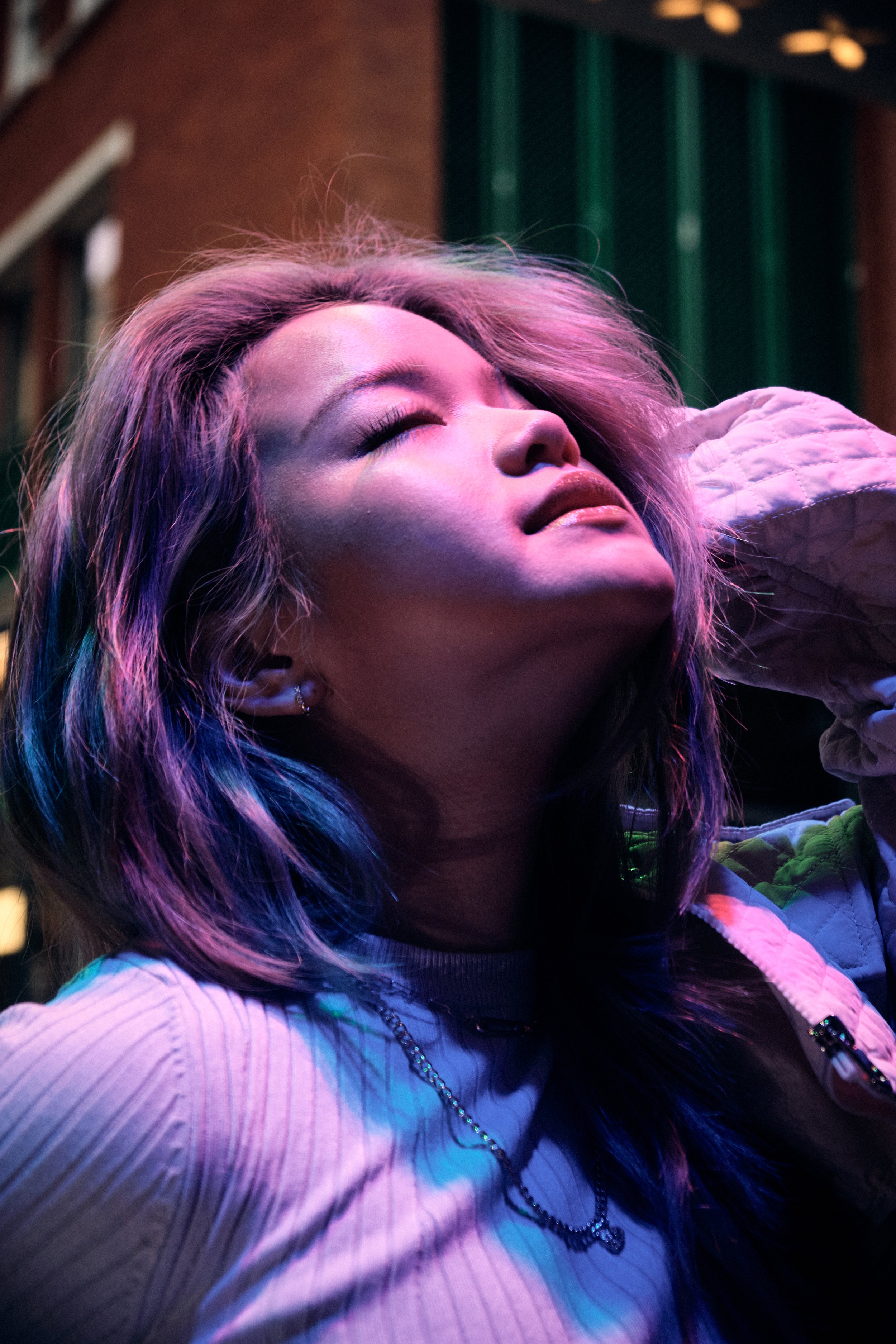 Person with colorful lighting on face, eyes closed, wearing a ribbed sweater and a necklace.