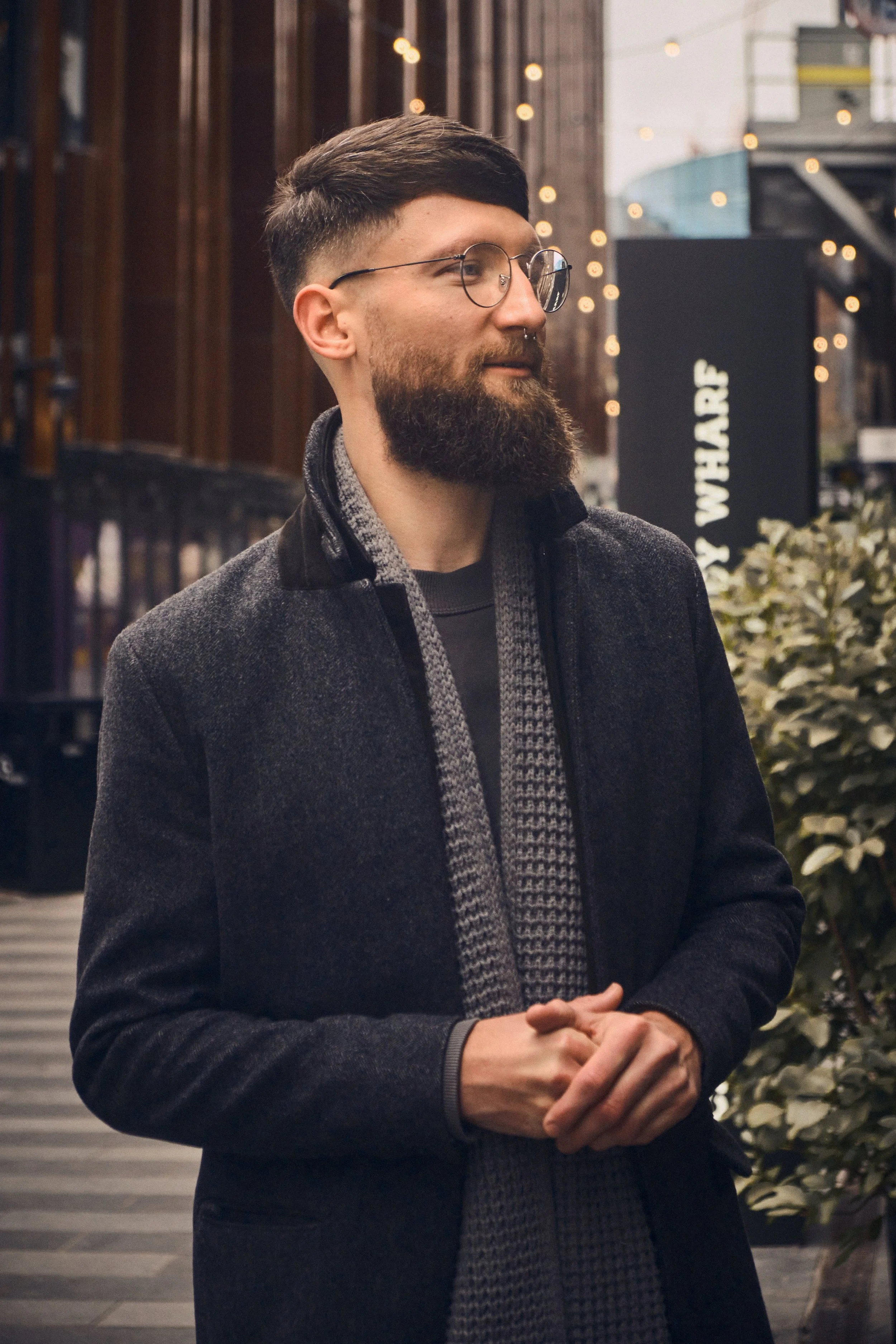 Man with glasses and beard standing outdoors, wearing a coat and scarf, with a blurred urban background.
