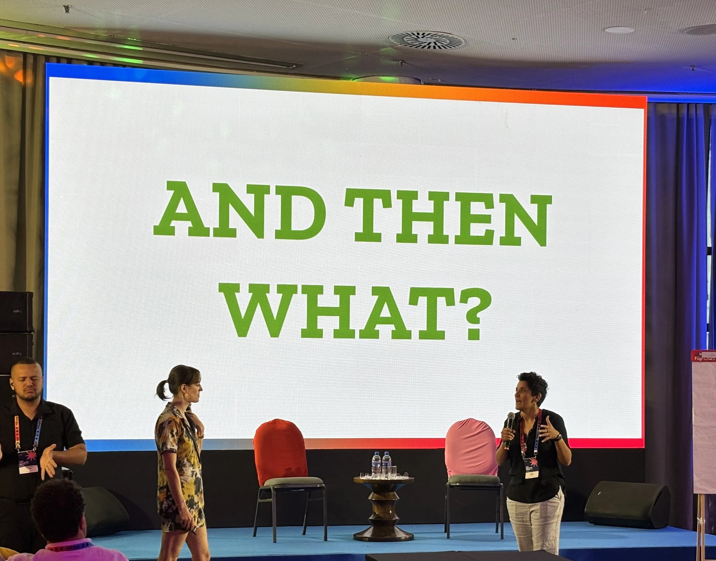 two artivism presenters with microphones in front of a screen that says "And Then What?" in big green letters on a white background