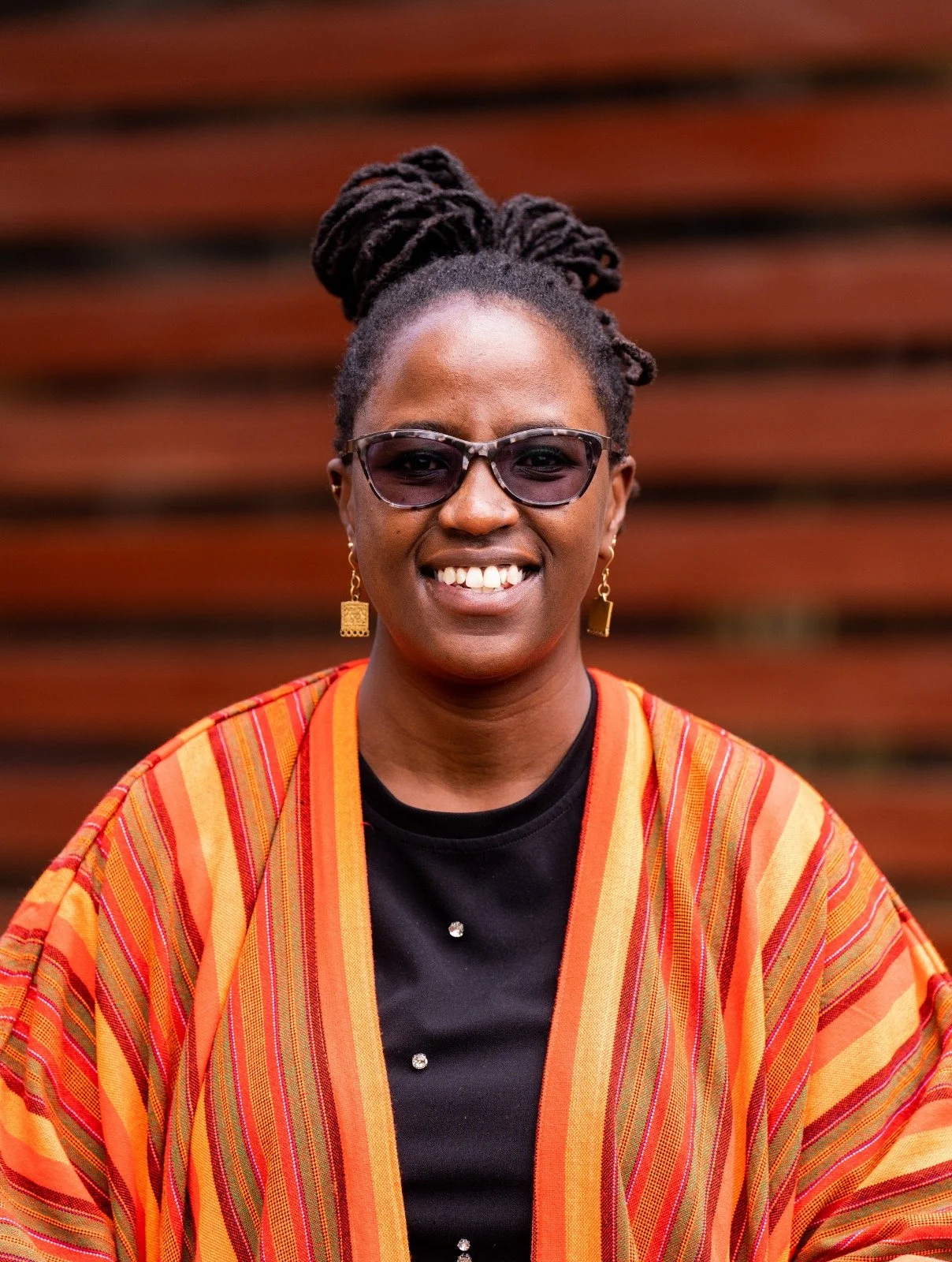 image of Susan Mbogo wearing a black top with a colorful cardigan, earrings, and sunglasses while smiling directly into the camera