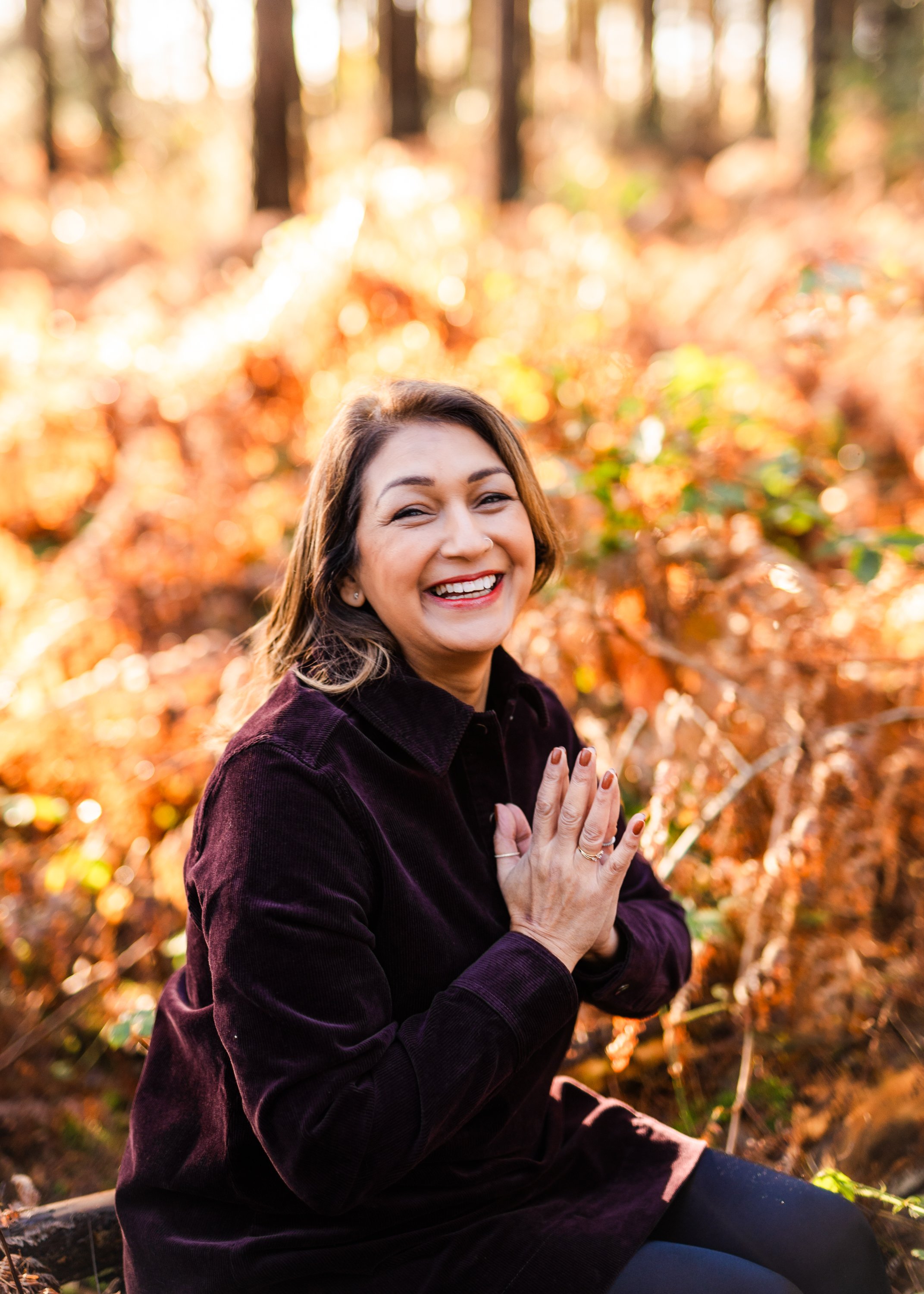 A woman smiling and with joined hands outdoors in a forest with autumn leaves.