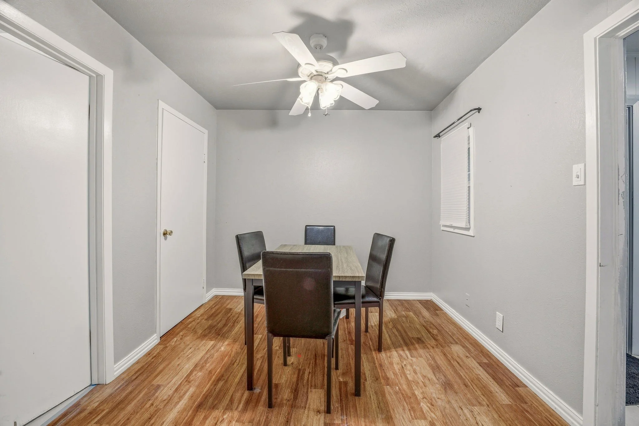A small dining area with a rectangular table and five dark-colored chairs, a white ceiling fan with light fixtures, white walls, one window with closed blinds, and a hardwood floor.