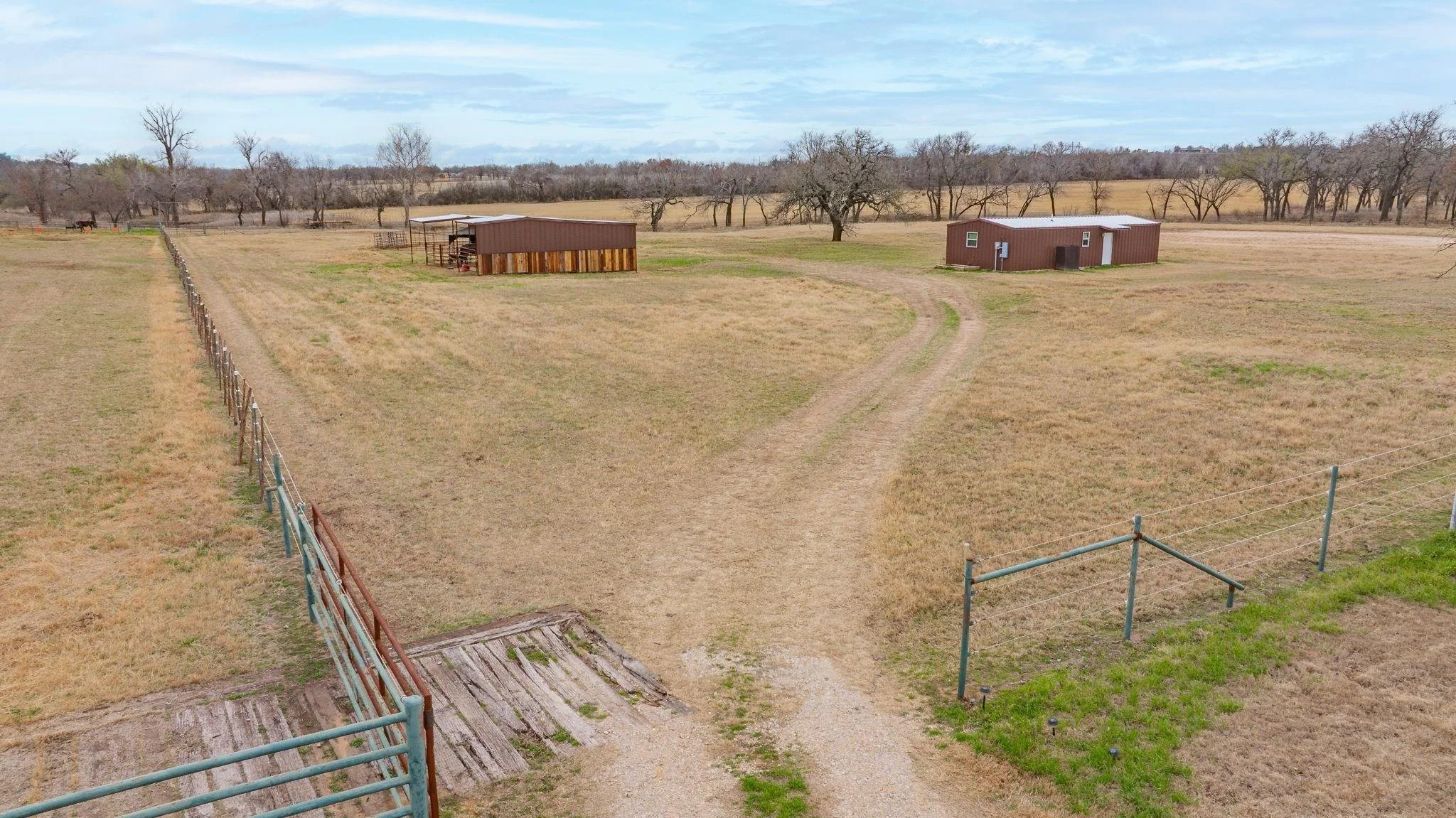 A rural farm landscape with dry grass, two small brown metal buildings, a dirt path leading to them, and trees in the background under a partly cloudy sky.