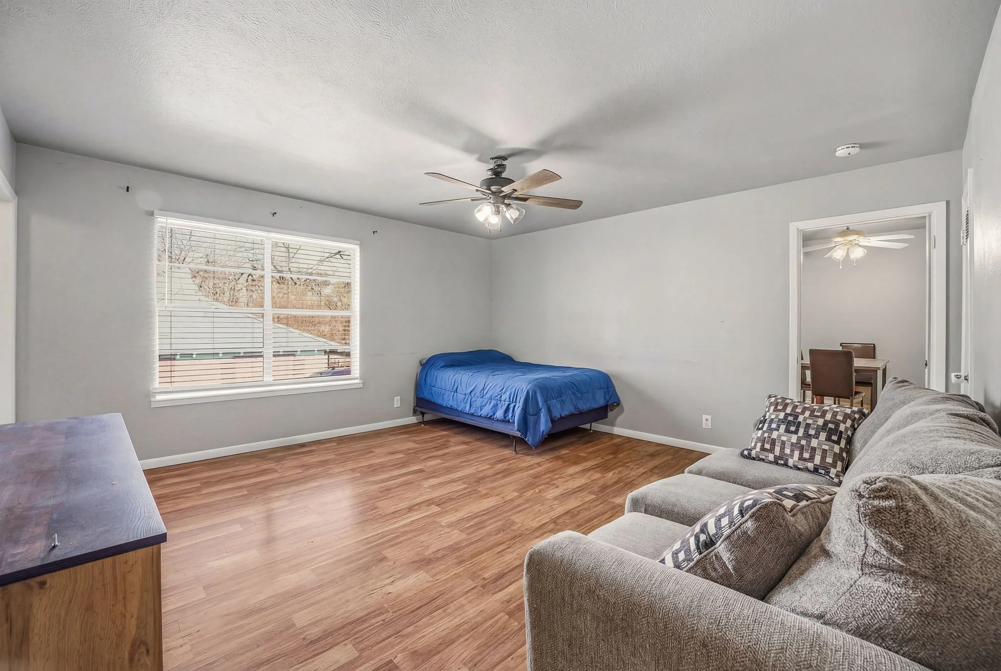 A spacious living room with a large window, a gray couch with pillows, a wooden side table, a bed with a blue comforter, and a ceiling fan. There is an adjacent dining area with a table and chairs visible through an open doorway.
