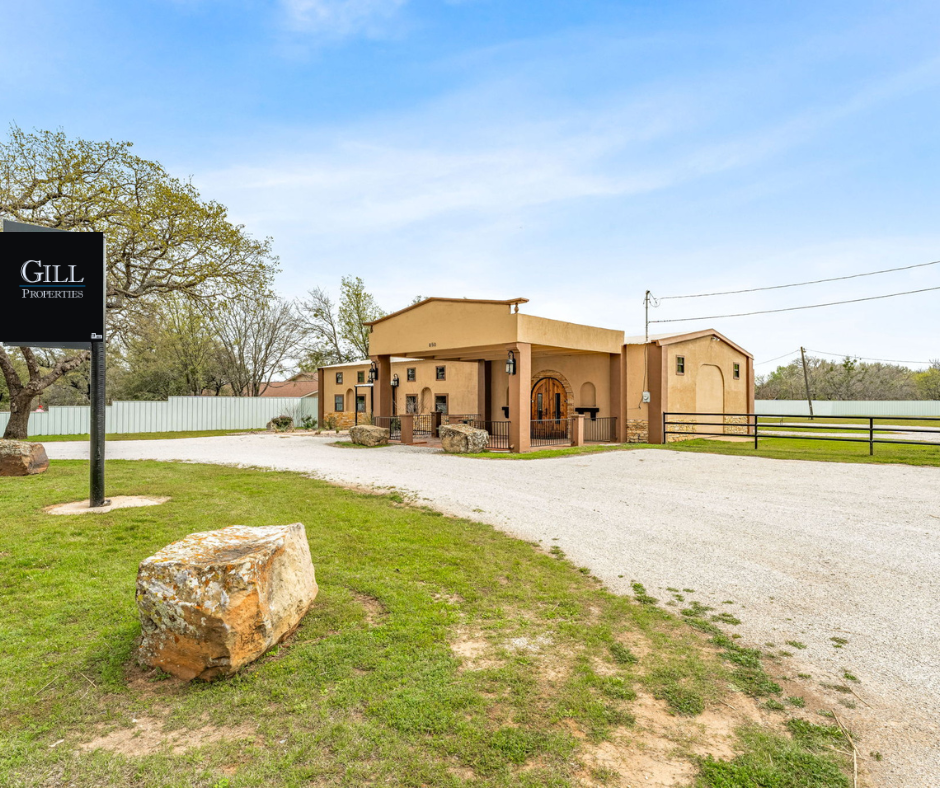 A building with a sign that reads 'Gill Properties' in a grassy area with large rocks, a gravel driveway, and trees behind a white fence.