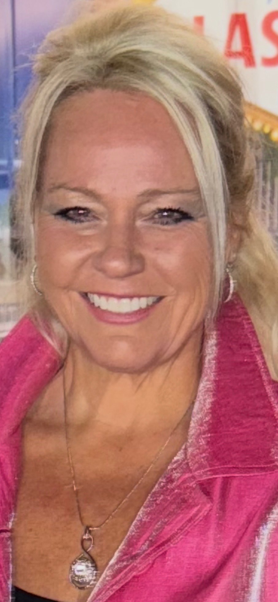 Close-up of a smiling woman with blonde hair, wearing a pink jacket, a silver necklace with a heart-shaped pendant, and earrings, in an indoor setting with a blurred background.