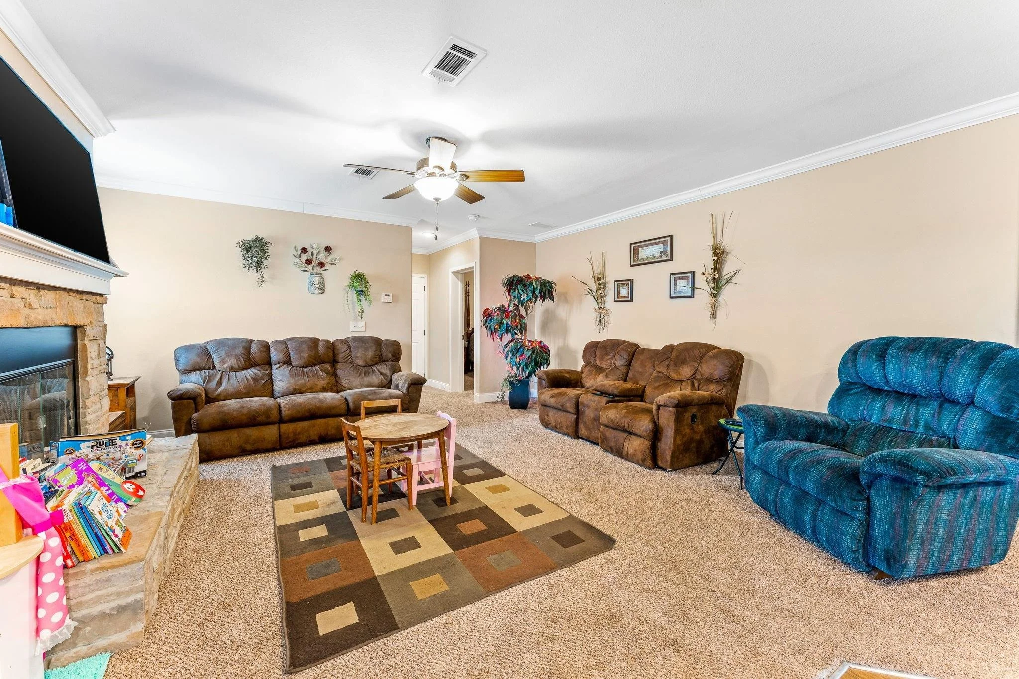 Living room with three couches, a rug, a small children's table, a fireplace, a television, wall decor, and a ceiling fan.