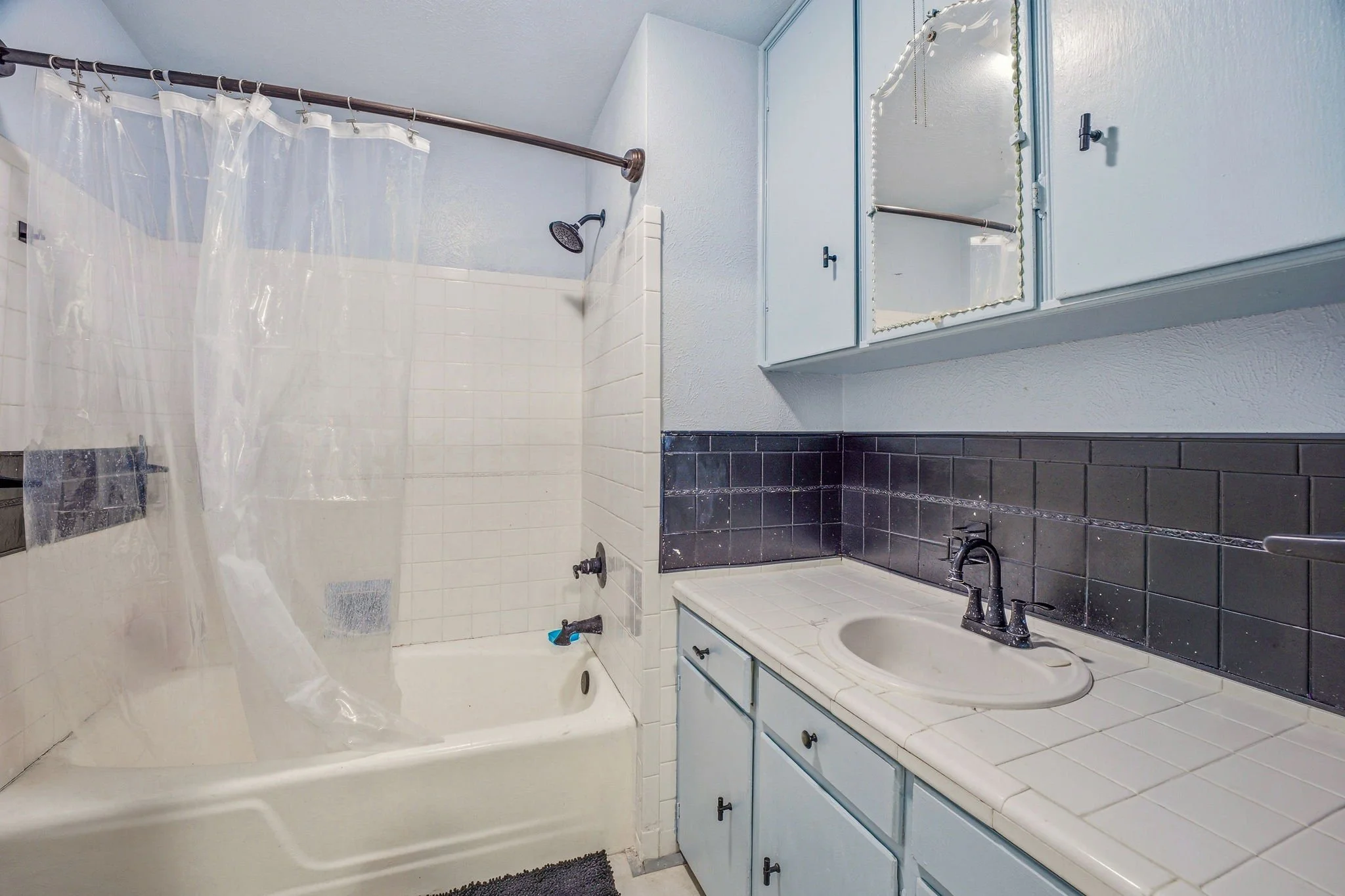 A bathroom with a bathtub and shower, white tiled walls, a dark gray tile backsplash above the sink, light blue cabinet and walls, and a mirror with decorative lights.