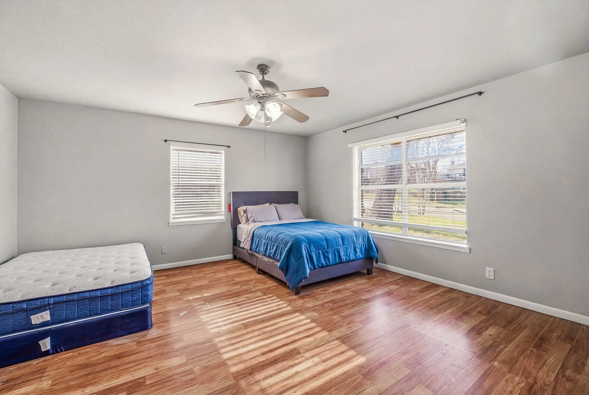 A bedroom with two windows, a ceiling fan, a bed with a blue comforter, and a mattress with no bed frame on the floor.