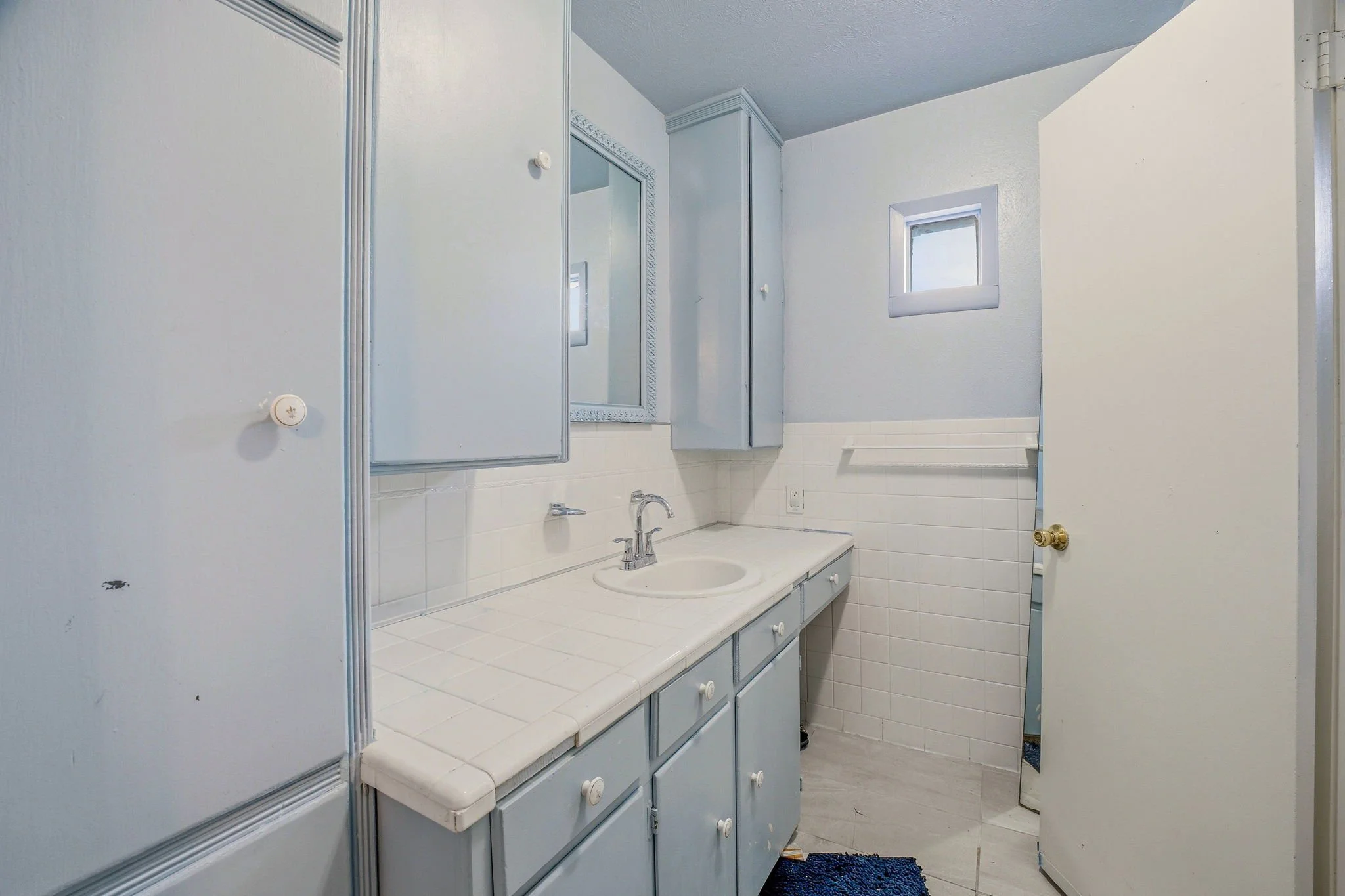 A small bathroom with a light blue vanity, white countertop, and a silver sink faucet. Above the sink is a mirror, and there are light blue cabinets above and beside the mirror. To the right, a small window is visible on the wall, with a white wall a
