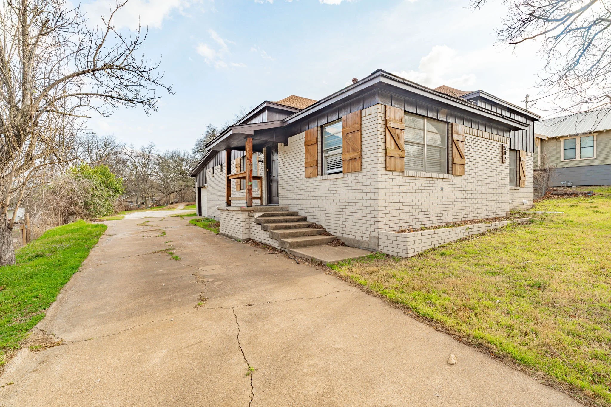 A brick house with a concrete front porch and steps, wooden shutters on the windows, and a driveway leading to the garage. Tree on the left with some greenery, and a grassy yard on the right.