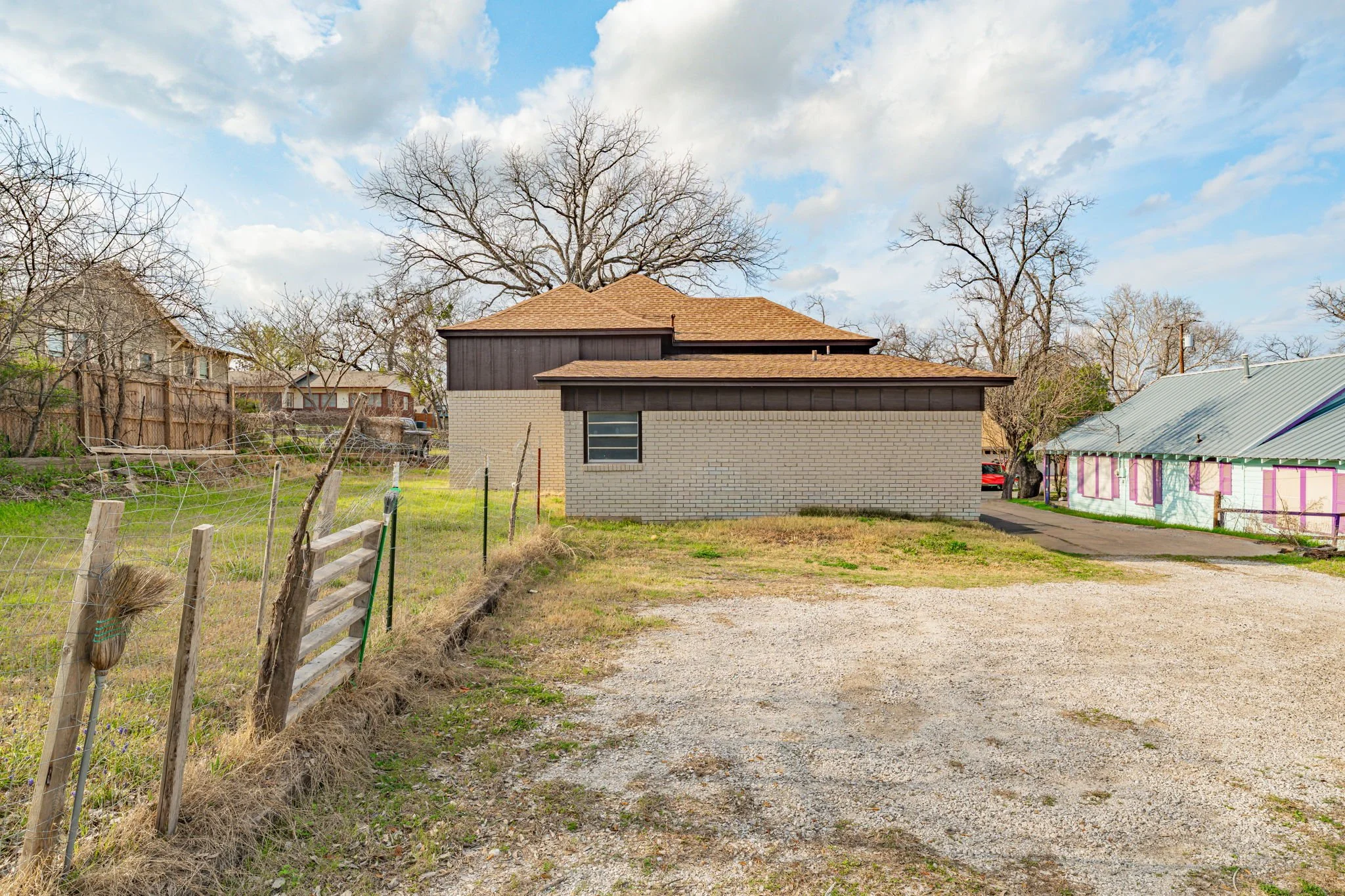 A residential backyard with a small brick house, a gravel driveway, an overgrown grassy area, a wooden fence on the left, and trees with leafless branches under a partly cloudy sky.