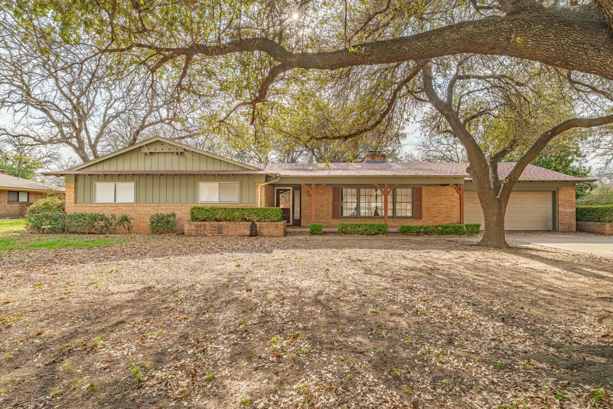 A single-story brick house with a front porch, large front window, attached garage, and a large tree in the front yard with bare branches and some green leaves.