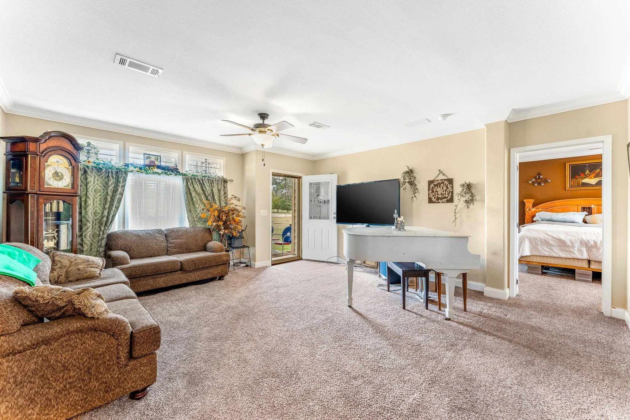 Living room with beige walls, a ceiling fan, a large flat-screen TV on a white grand piano, brown sofas, an antique clock, green patterned curtains, and a door leading outside. A doorway opens to a bedroom with a wooden bed frame.