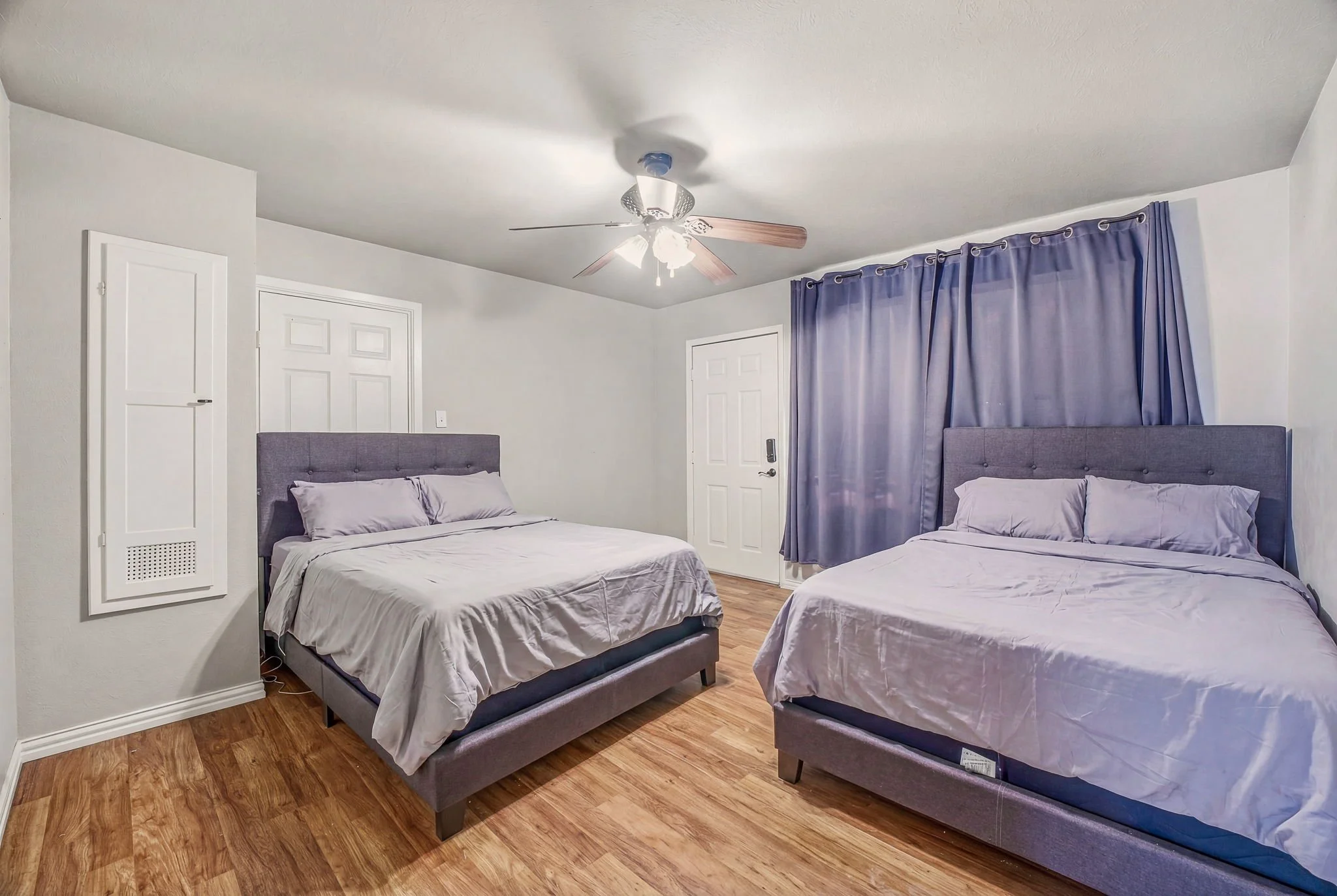 Two beds in a bedroom with purple headboards, gray bedding, purple curtains, wood flooring, a ceiling fan, and white walls.