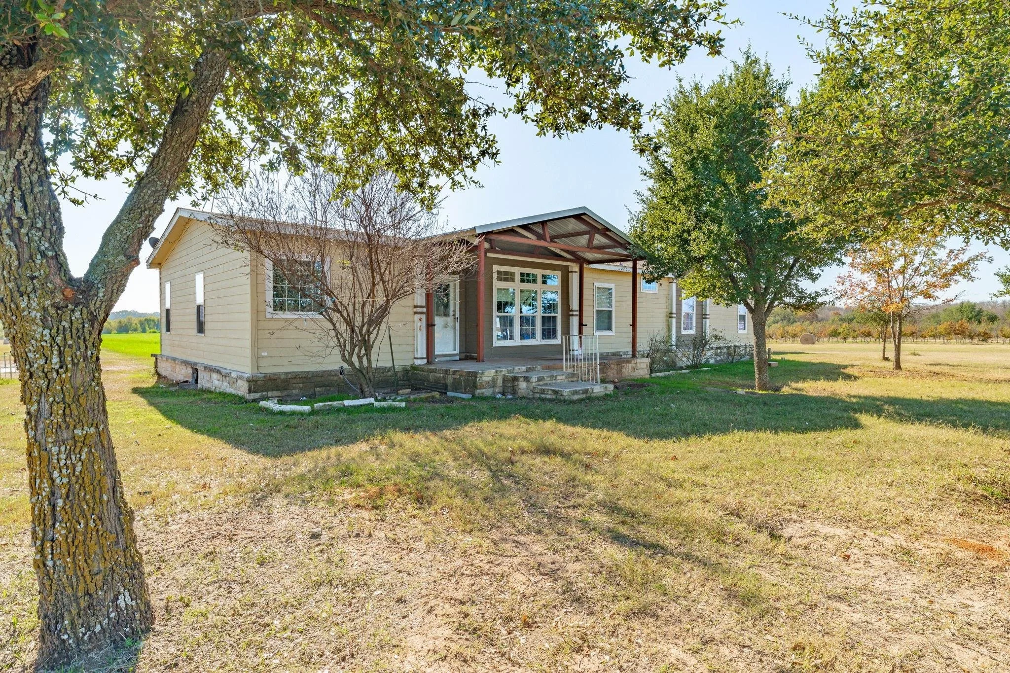 A single-story house with light yellow siding, a small porch with steps, and surrounded by a grassy yard with trees, under a clear blue sky.