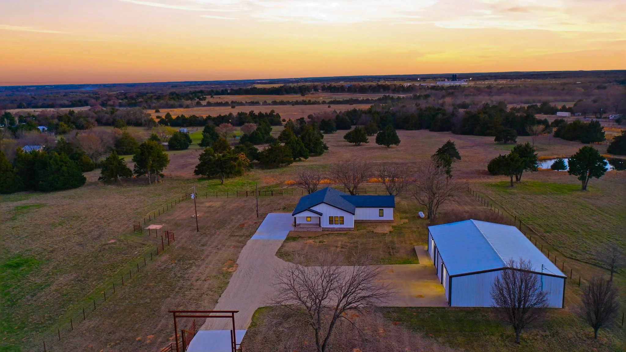 Aerial view of a rural property at sunset with a house, a large garage, trees, fenced areas, and nearby ponds in the distance.