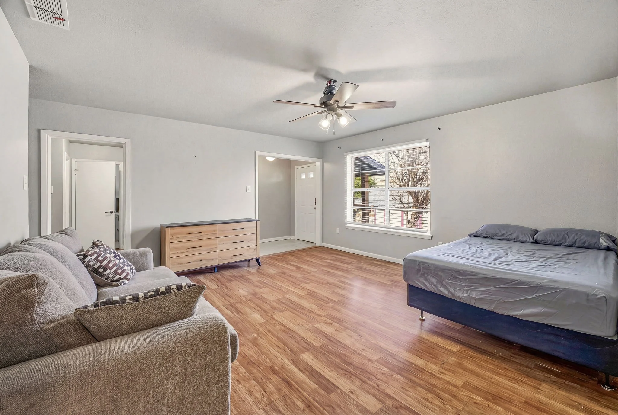 Living room with a beige sofa, a wooden dresser, a bed with gray bedding, large window, and hardwood flooring.