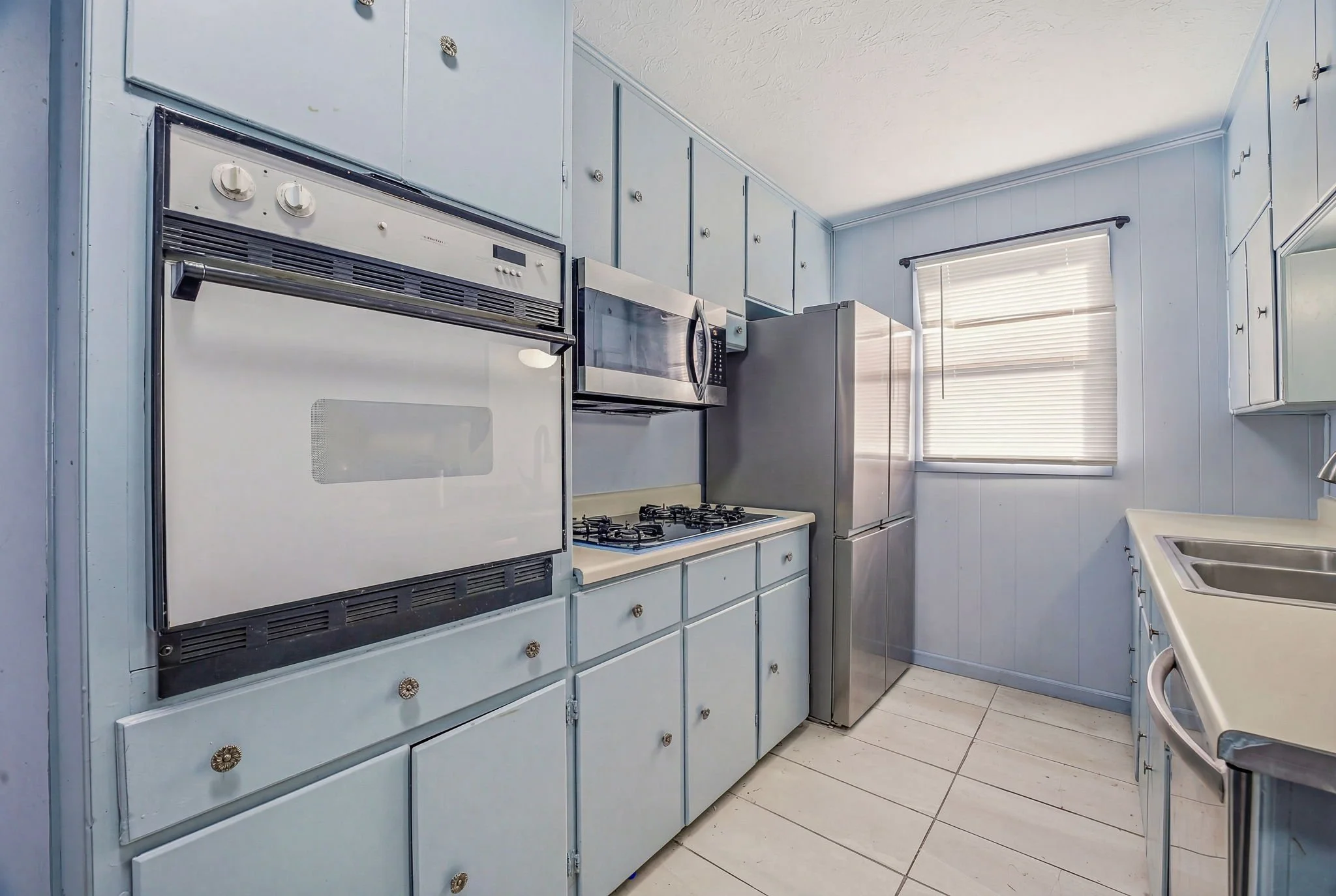 A small kitchen with light blue cabinets, a white countertop, a stainless steel refrigerator, a microwave oven, a gas stove, a double sink, and a window with white blinds.