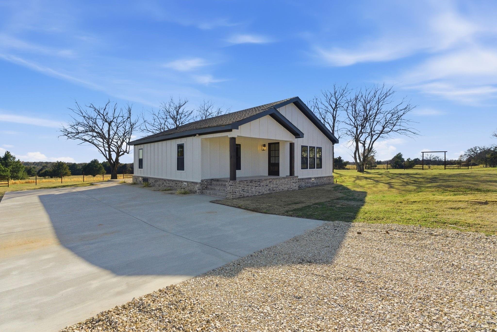 A newly built small white house with dark trim, a front porch, and brick foundation, situated on a spacious grassy lot with a gravel and concrete driveway, leafless trees, and a blue sky with some clouds.