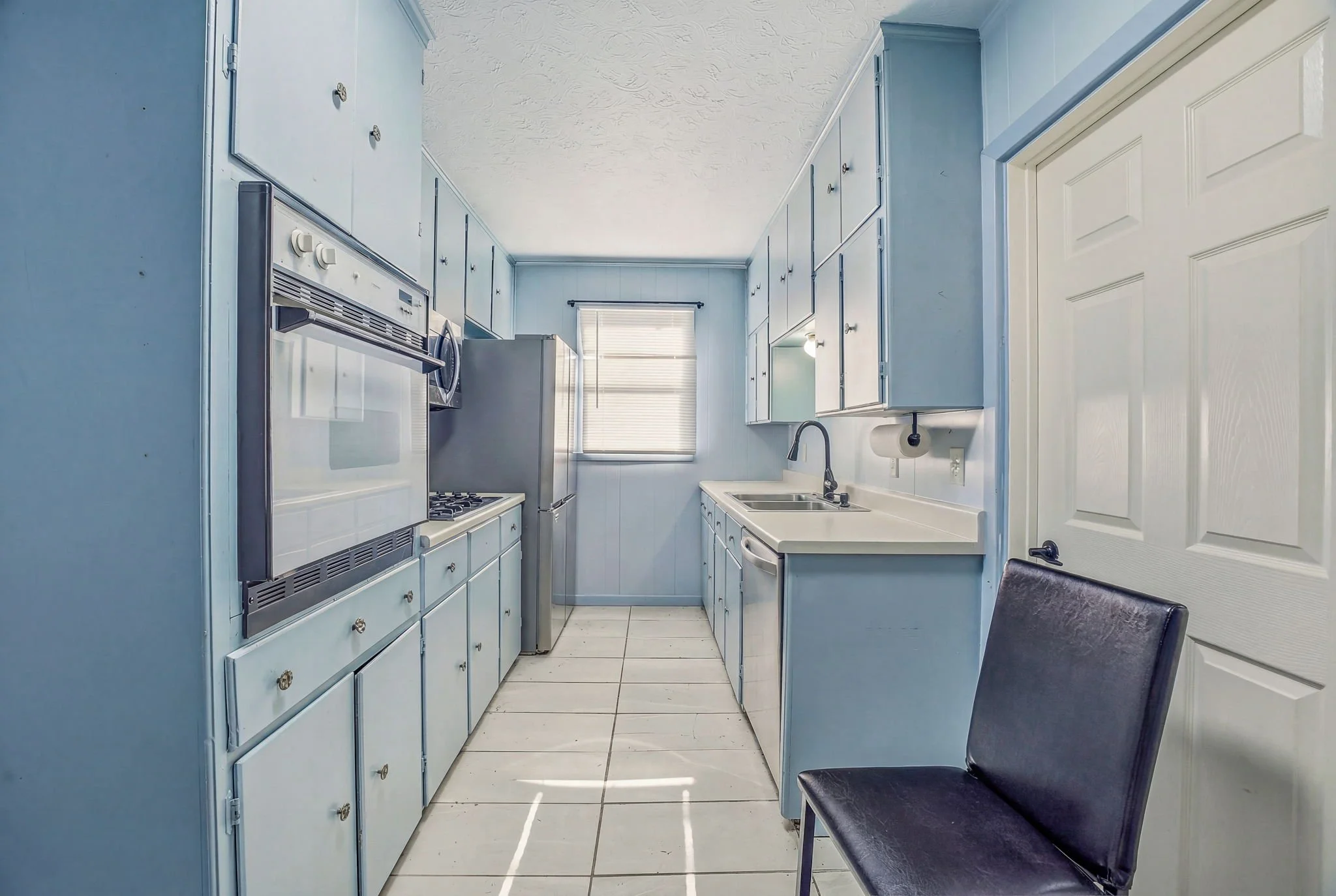 Small kitchen with light blue cabinets, a window, and a black chair in the foreground.
