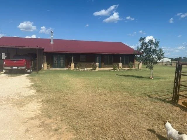 A house with a red metal roof and brick columns on a grassy lot, with a car parked under a carport, a small tree, and a dog in the yard. Blue sky with scattered clouds.