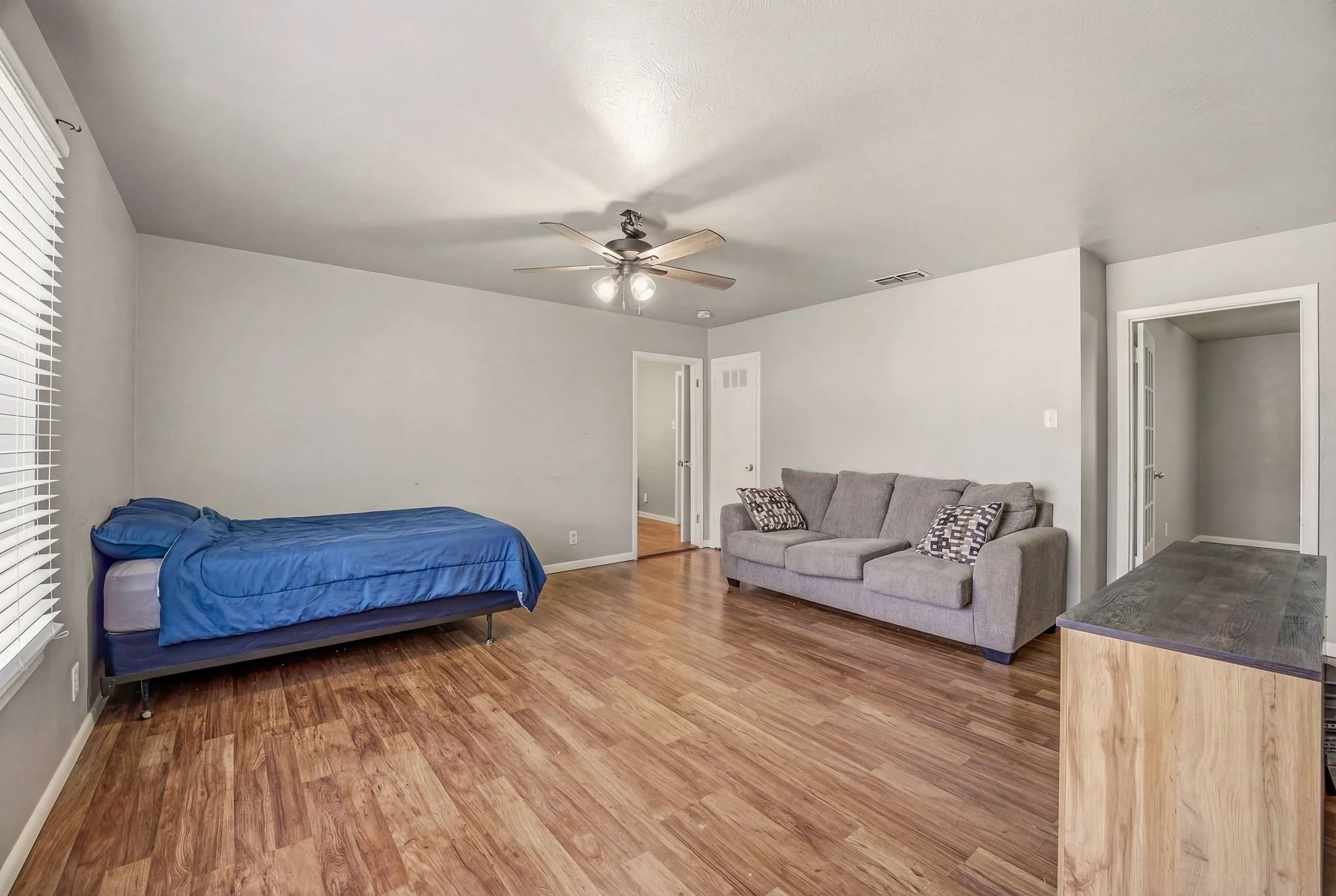 Living room with hardwood flooring, a single bed with blue bedding, a gray sofa with patterned pillows, a ceiling fan with lights, and a wooden TV stand.