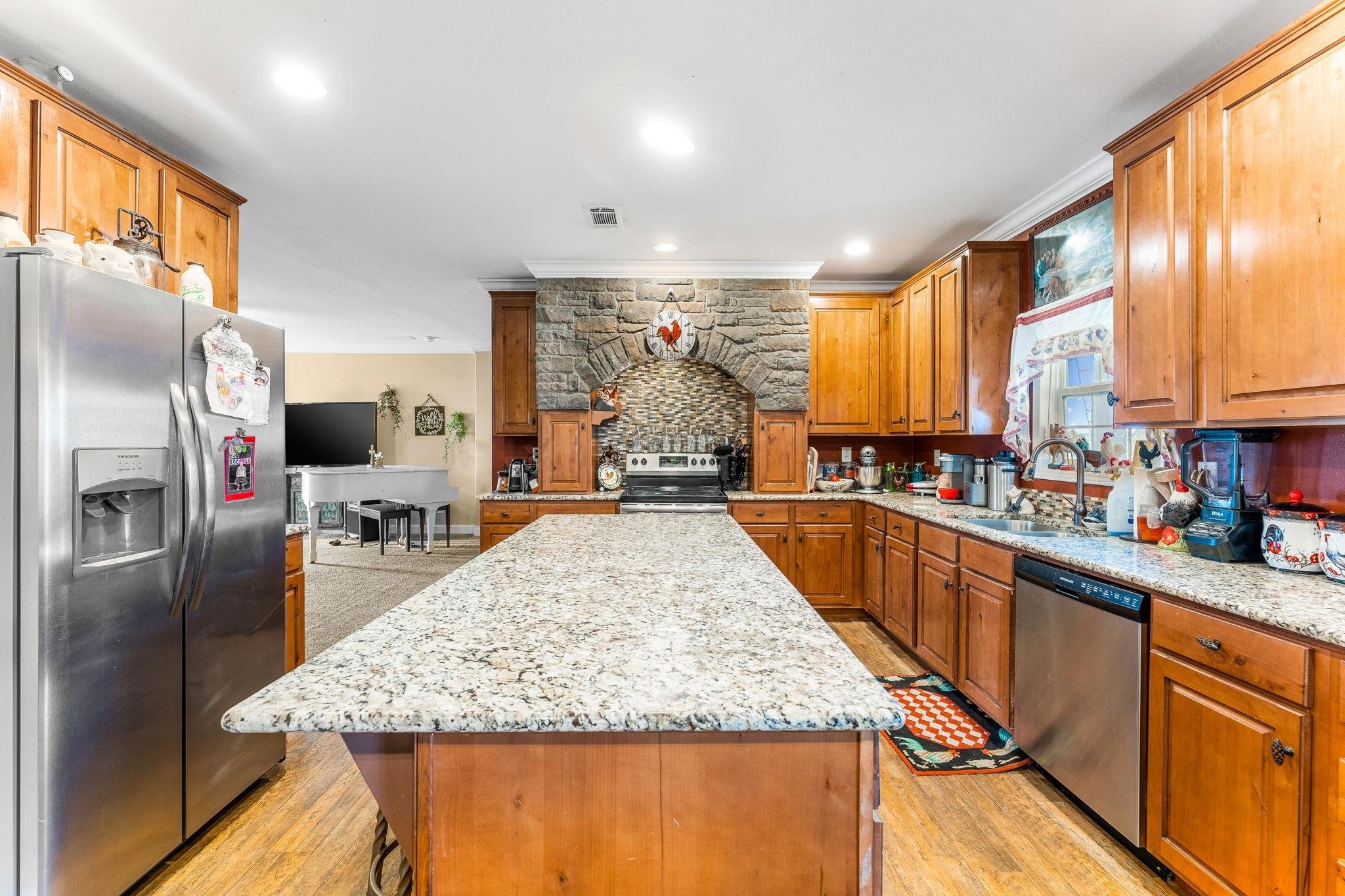 A spacious kitchen with wooden cabinets, granite countertops, stainless steel appliances, and a central island. The background features a brick and stone arch over the stove, with decorative pumpkin and rooster wall hangings. There is a window with a