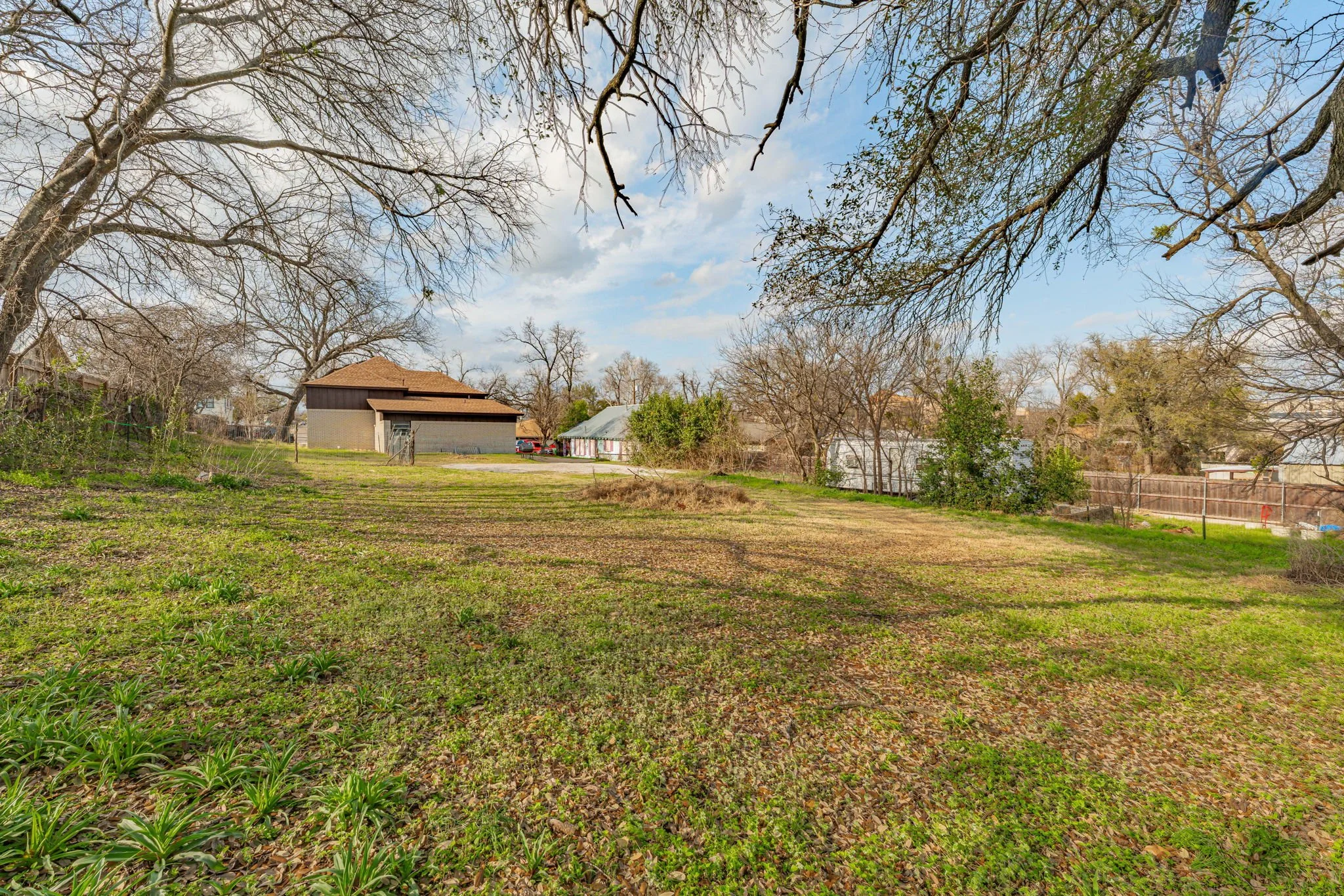 A backyard with bare trees, green grass, and patches of brown soil, with houses visible in the background under a partly cloudy sky.