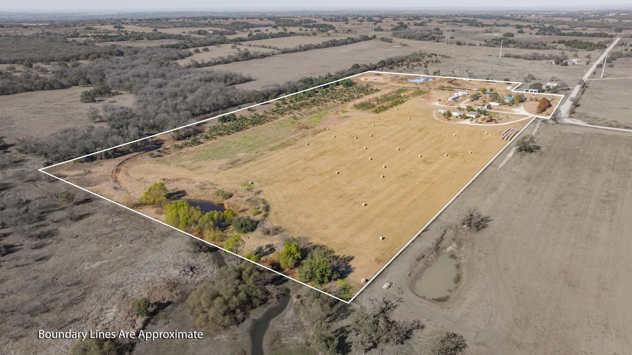 Aerial view of a large rural property with a mixture of open fields, trees, and a small pond, outlined by boundary lines, with a farmstead or residential area in the upper right corner.