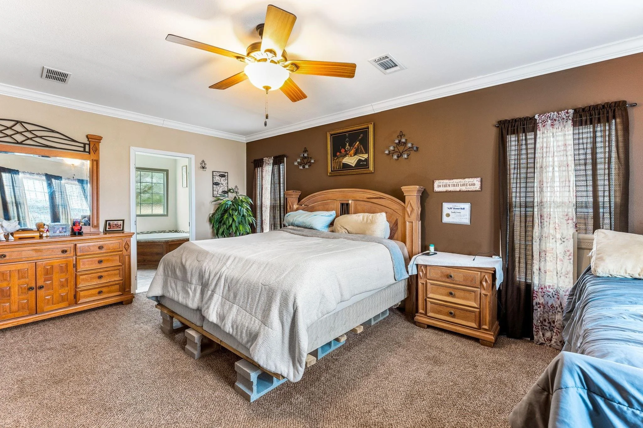 Bedroom with two beds, one on cinder blocks and the other on the floor, wooden nightstand, dresser with mirror, windows with curtains, ceiling fan, wall art, a potted plant, and framed certificates.