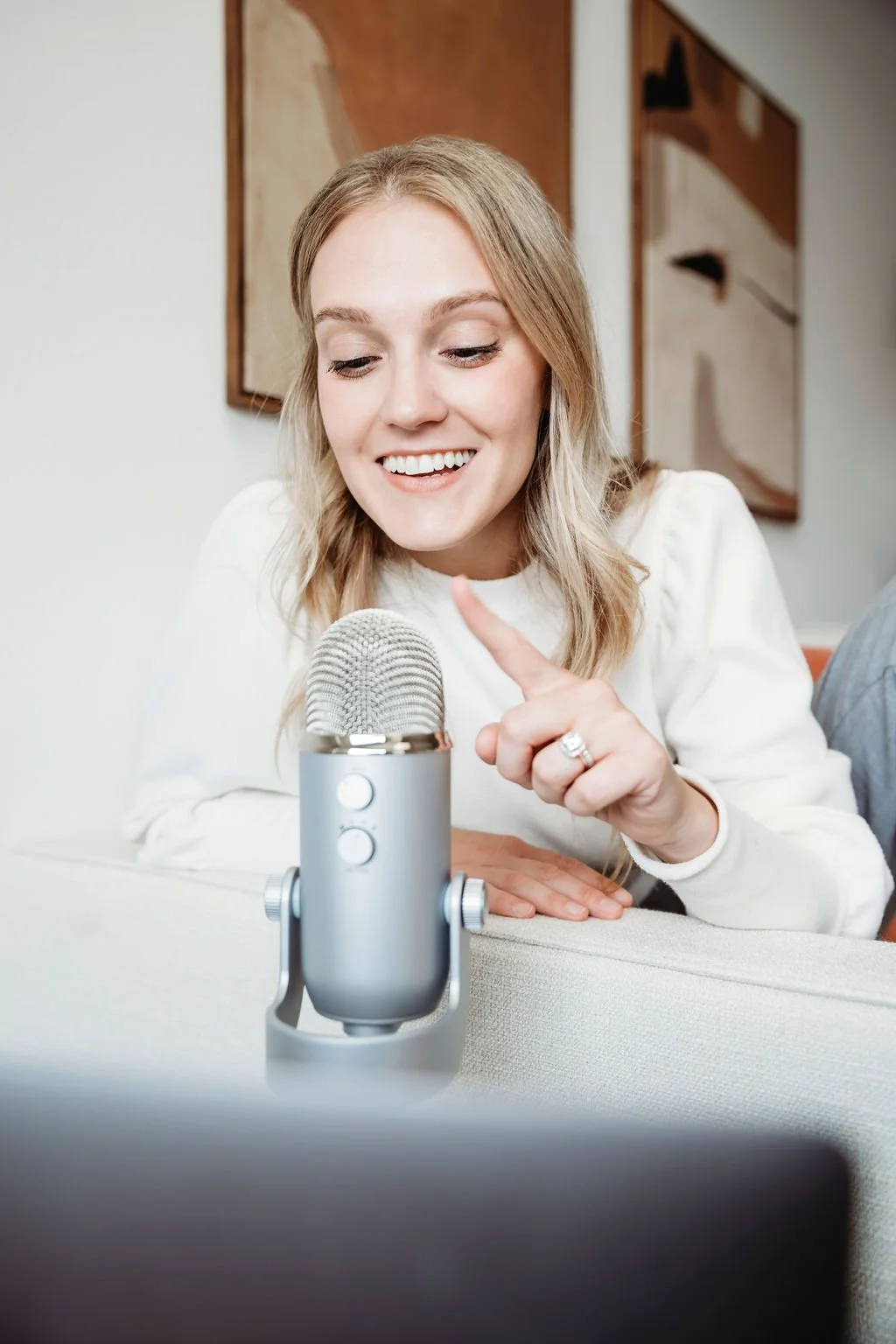 A woman smiling and pointing at a silver microphone while sitting on a couch.