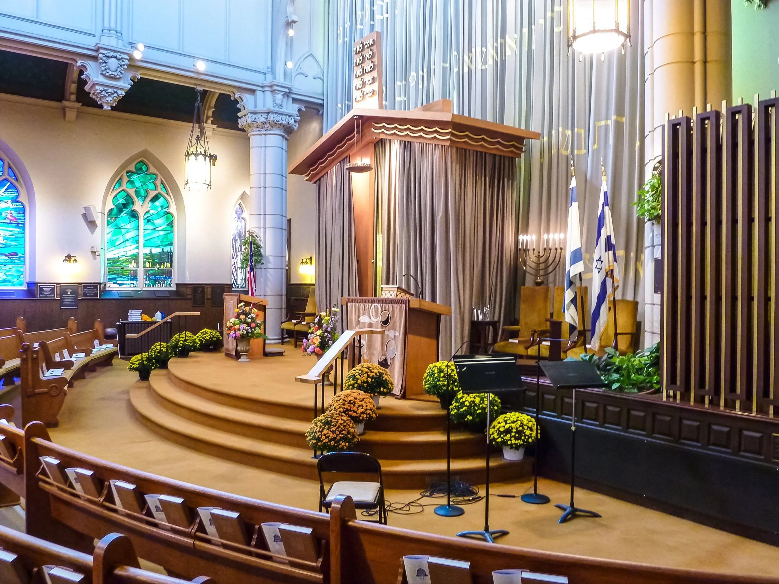 Interior of a religious sanctuary with a raised platform, floral arrangements, flags, stained glass windows, wooden pews, and artifacts for a Jewish service.