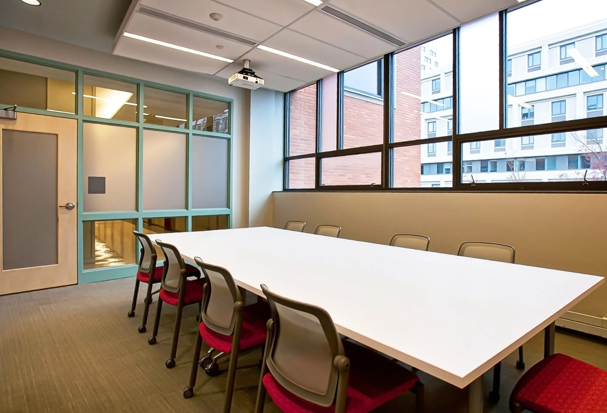 Empty conference room with a long white table, eight chairs with red cushions around it, large windows with a view of adjacent buildings, a ceiling projector, and frosted glass partition with a door.